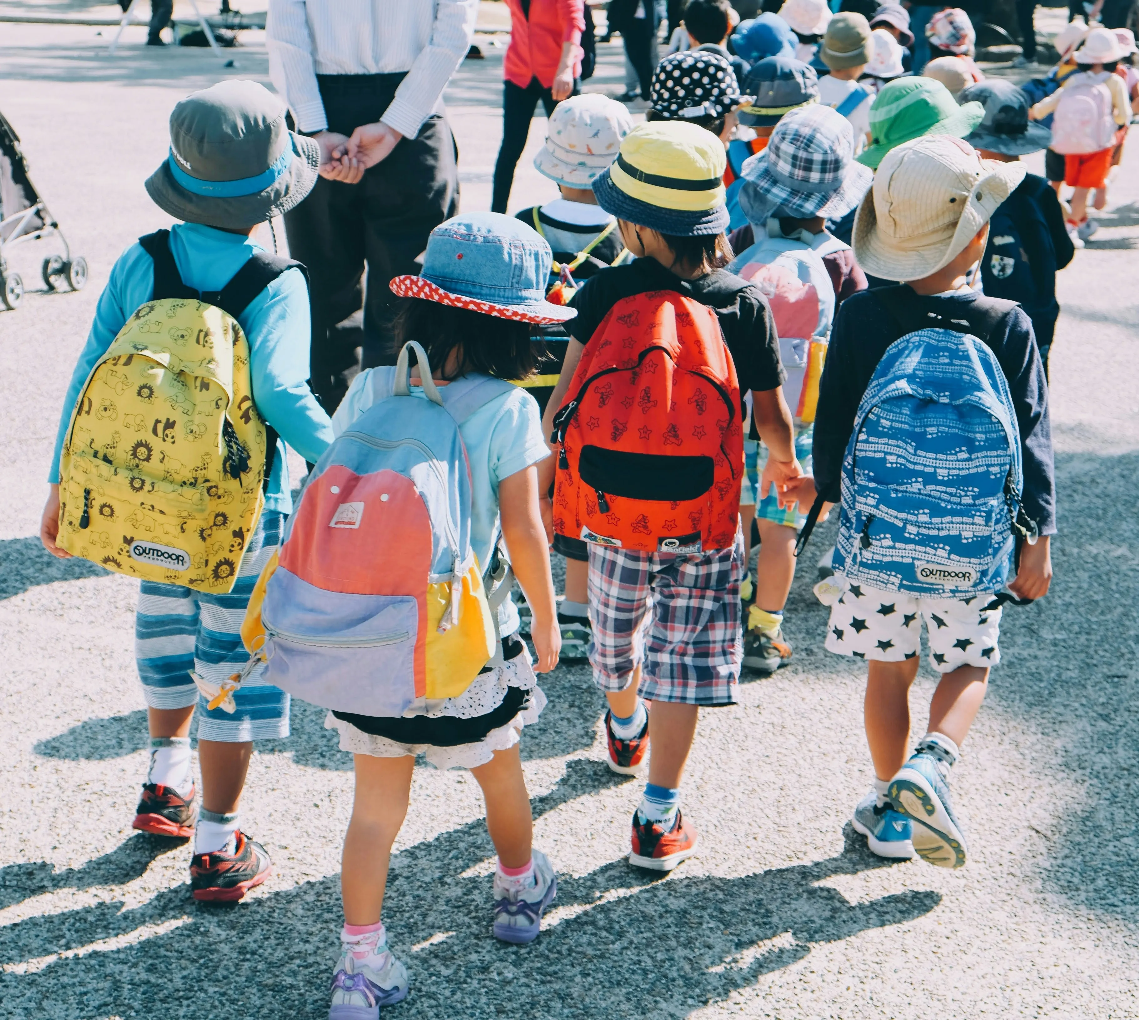 Group of elementary children wearing backpacks and hats walking outdoors on a sunny day.