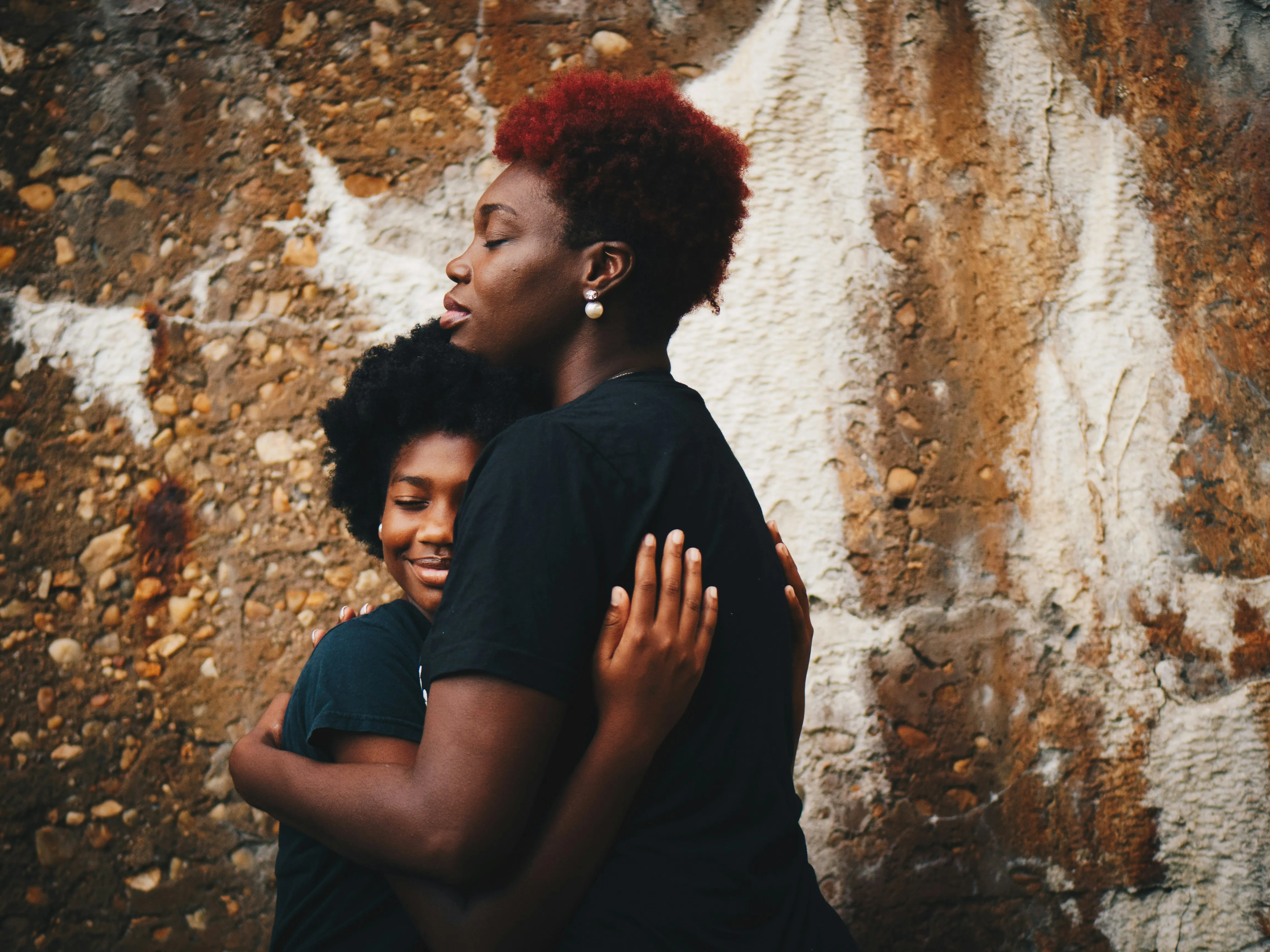 Two Black girls with natural hair embracing each other against a textured stone wall.