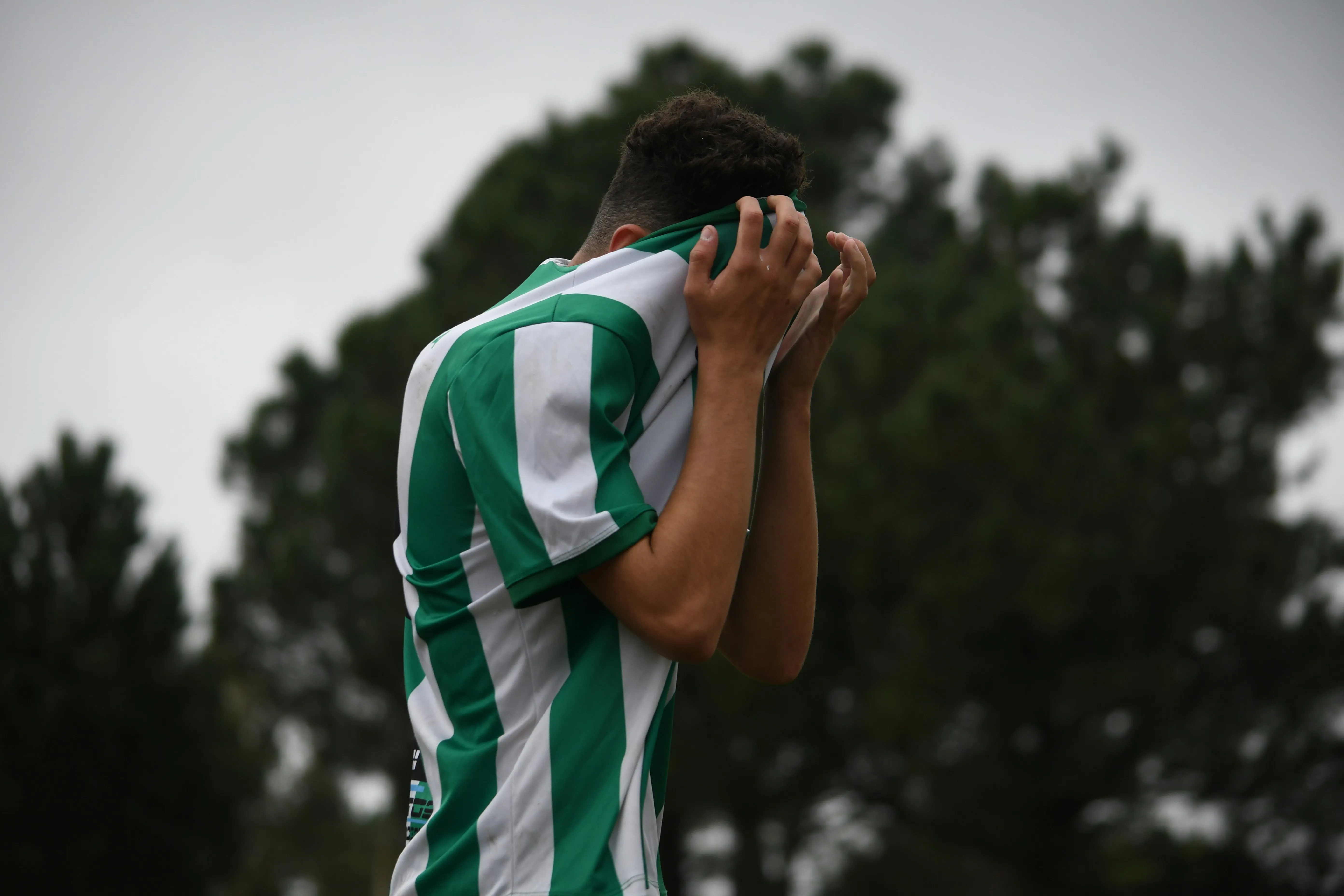 Soccer player in green and white jersey covering face with shirt outdoors near trees.