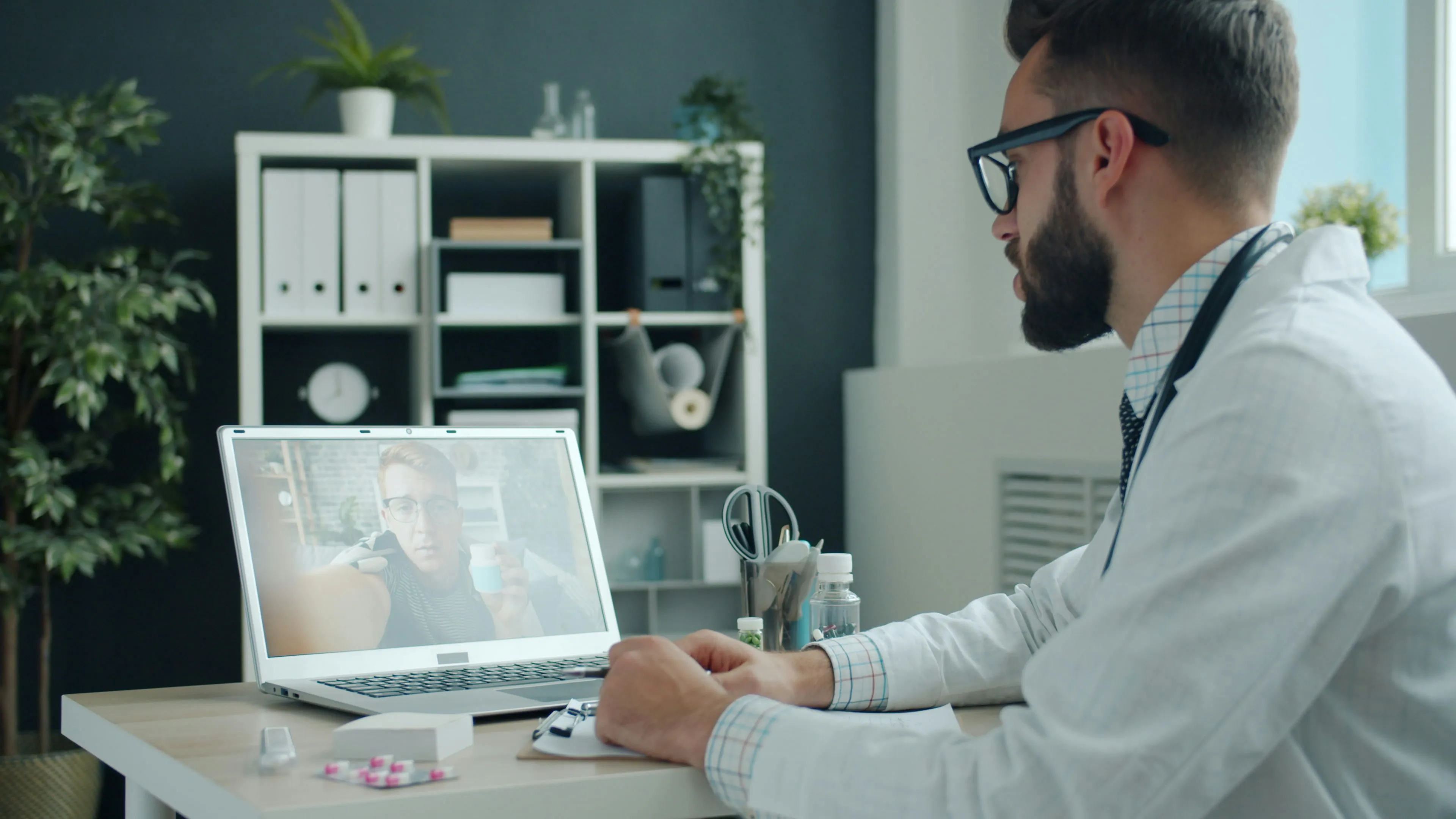 Doctor in white coat conducting a telehealth video call with a patient holding medication on laptop screen.