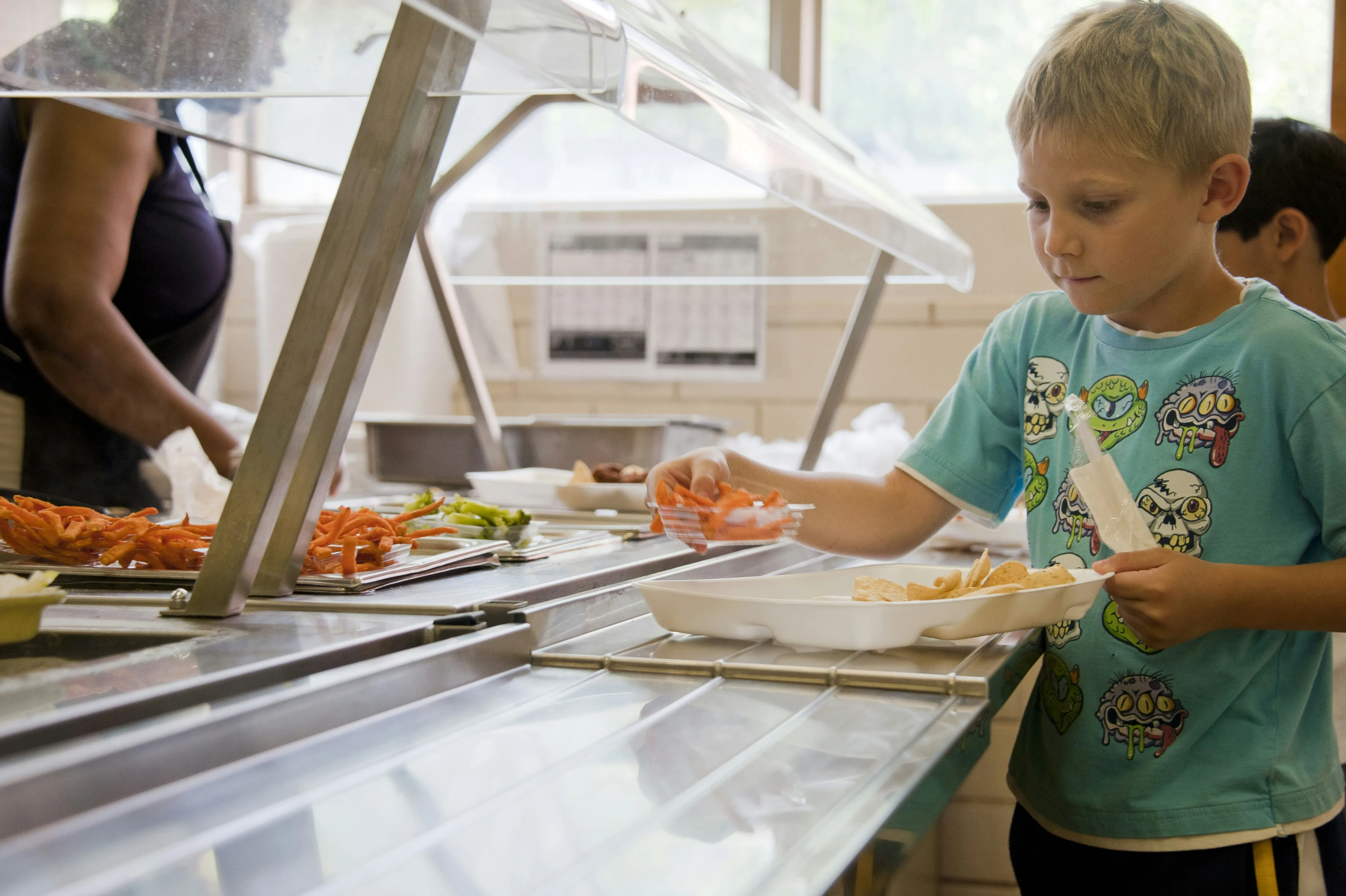 Young boy with light hair selecting carrots from a cafeteria line holding a tray with chips and a drink.