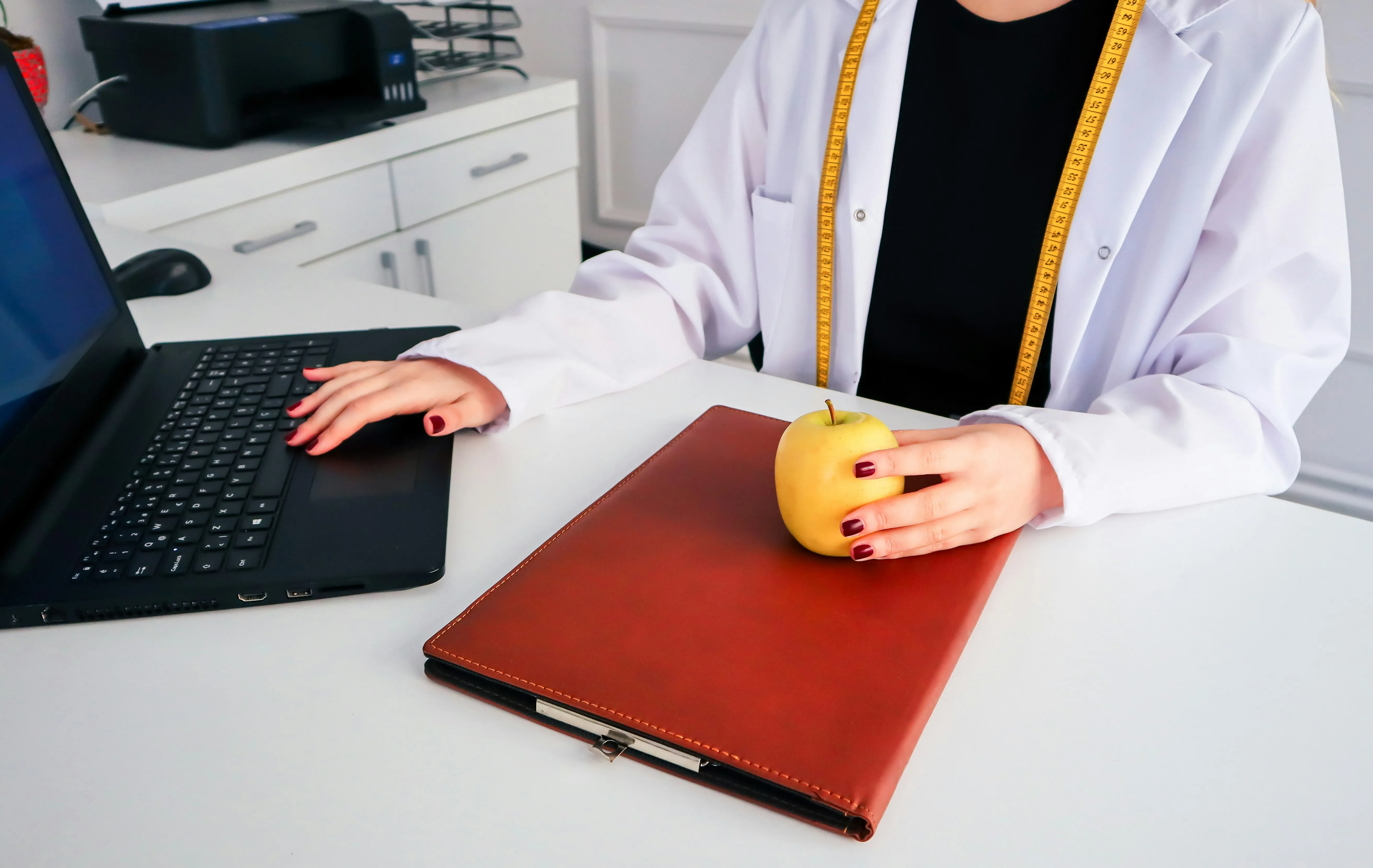 Person in white coat with measuring tape holds yellow apple near brown clipboard and laptop keyboard.