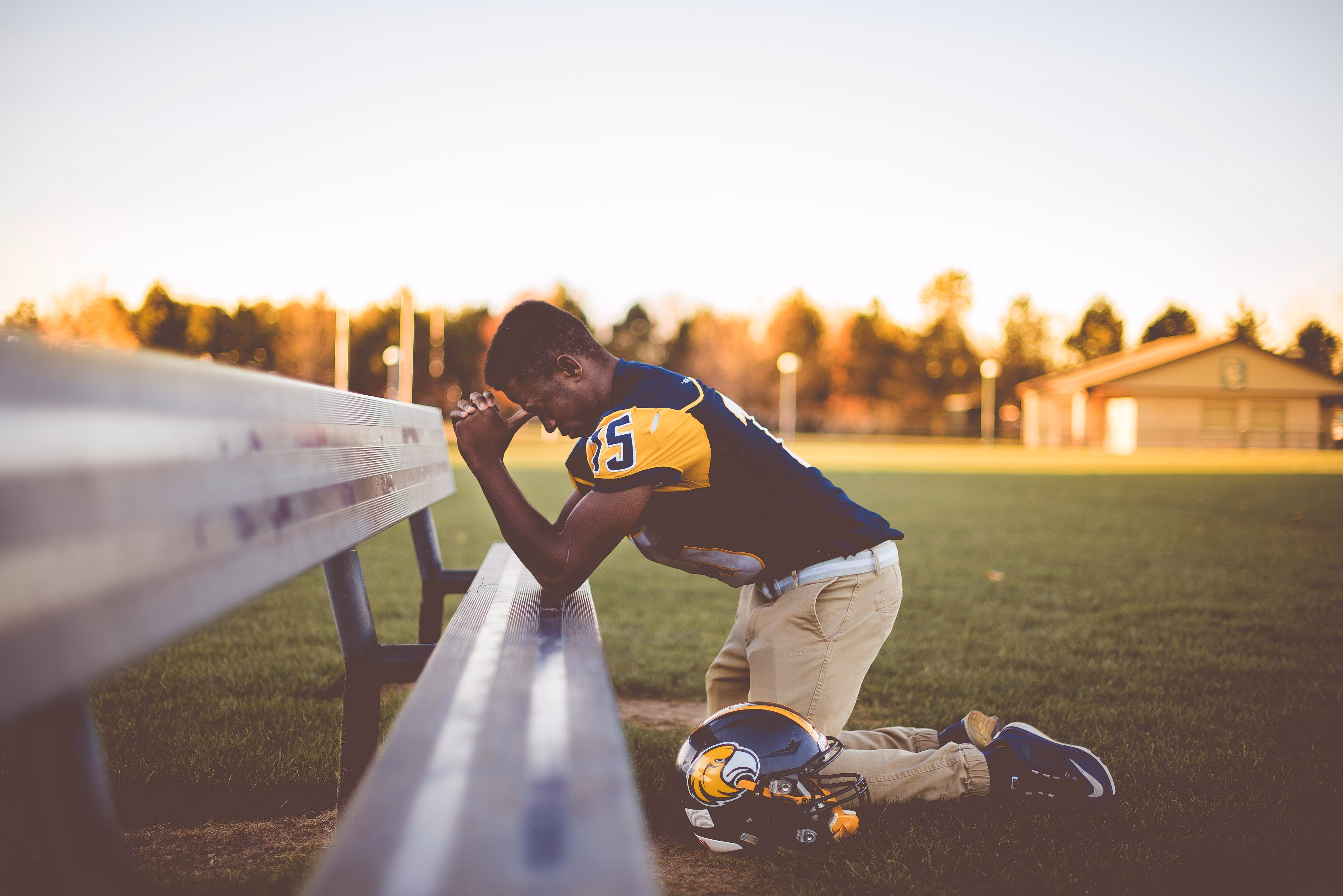 Young football player kneeling by a bench with helmet on grass, head bowed in reflection.