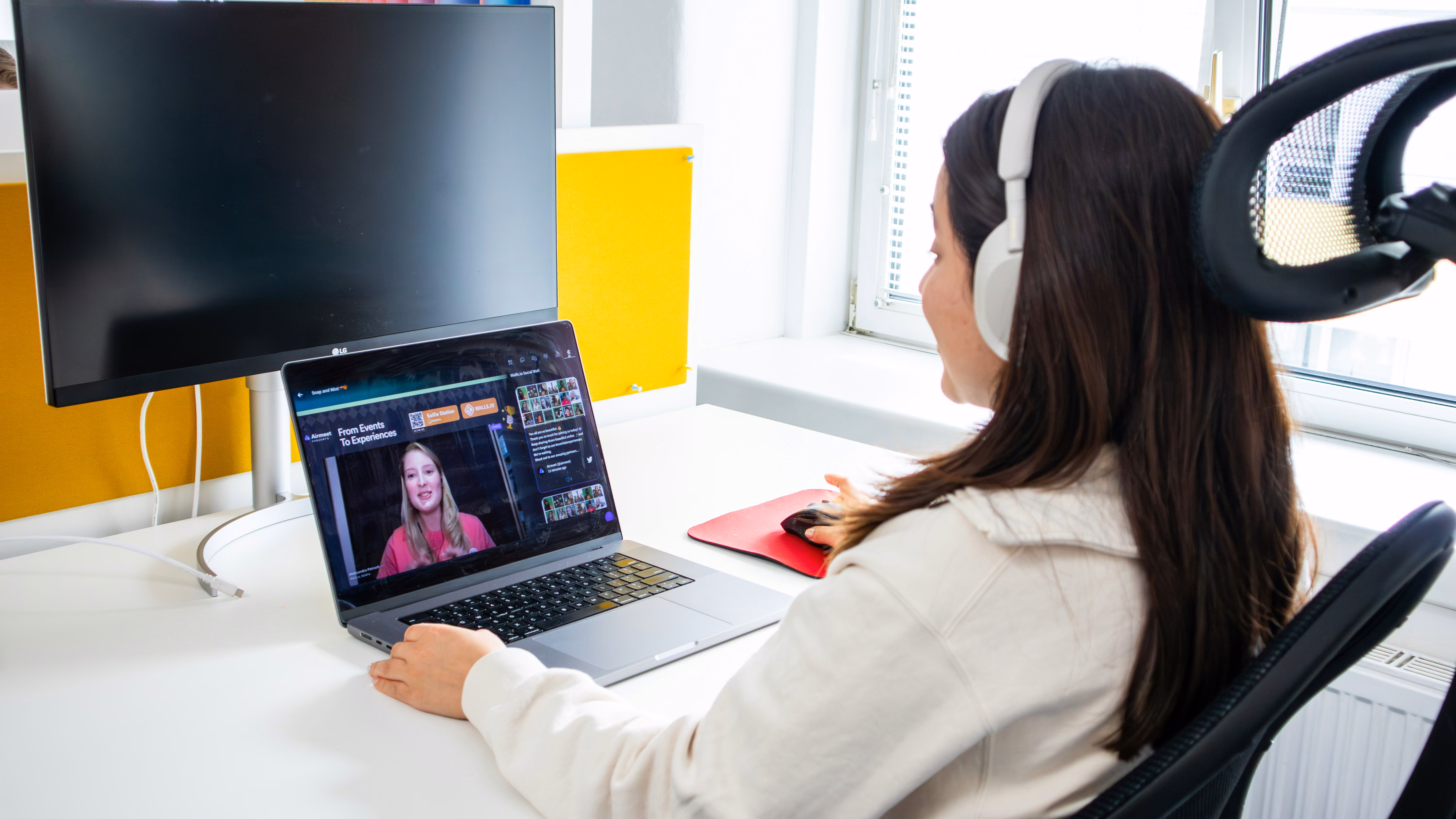 Woman with white headphones watches a virtual event on a laptop at a desk with a large monitor nearby.