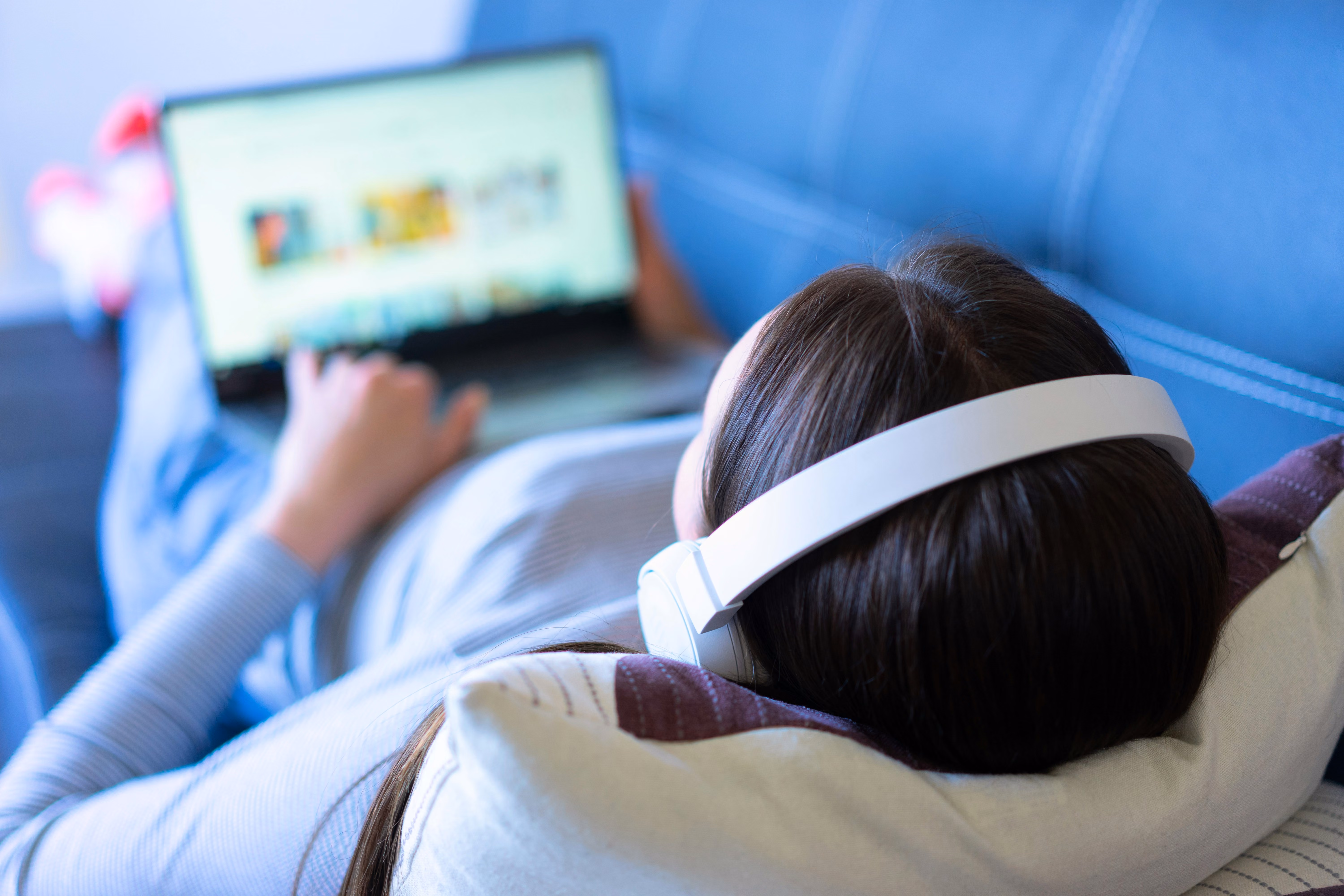 Person wearing white headphones reclining on a couch using a laptop with a blurry screen.