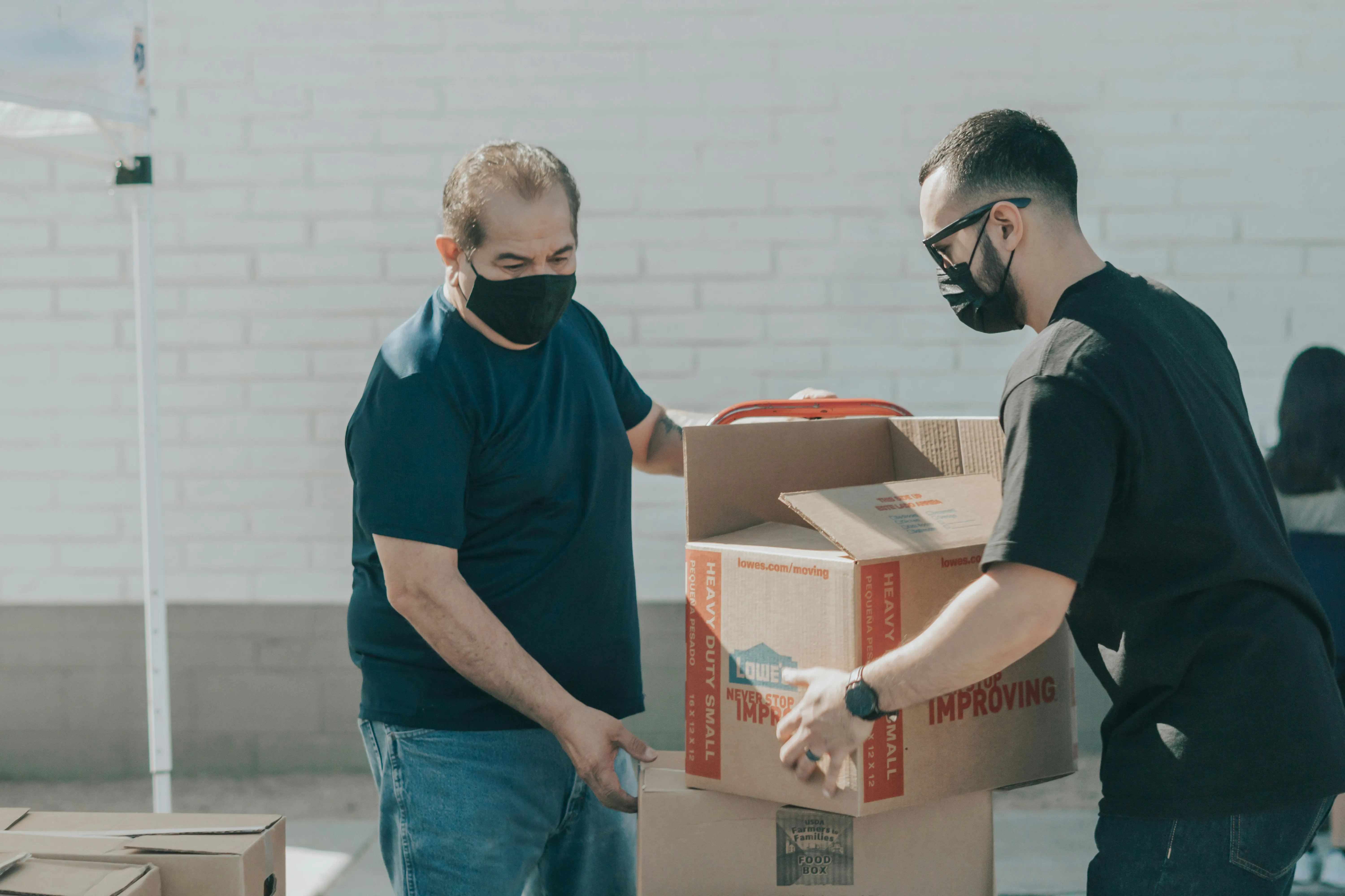 Two men wearing masks handle cardboard boxes outdoors, one labeled Food Box and Lowe's moving box.