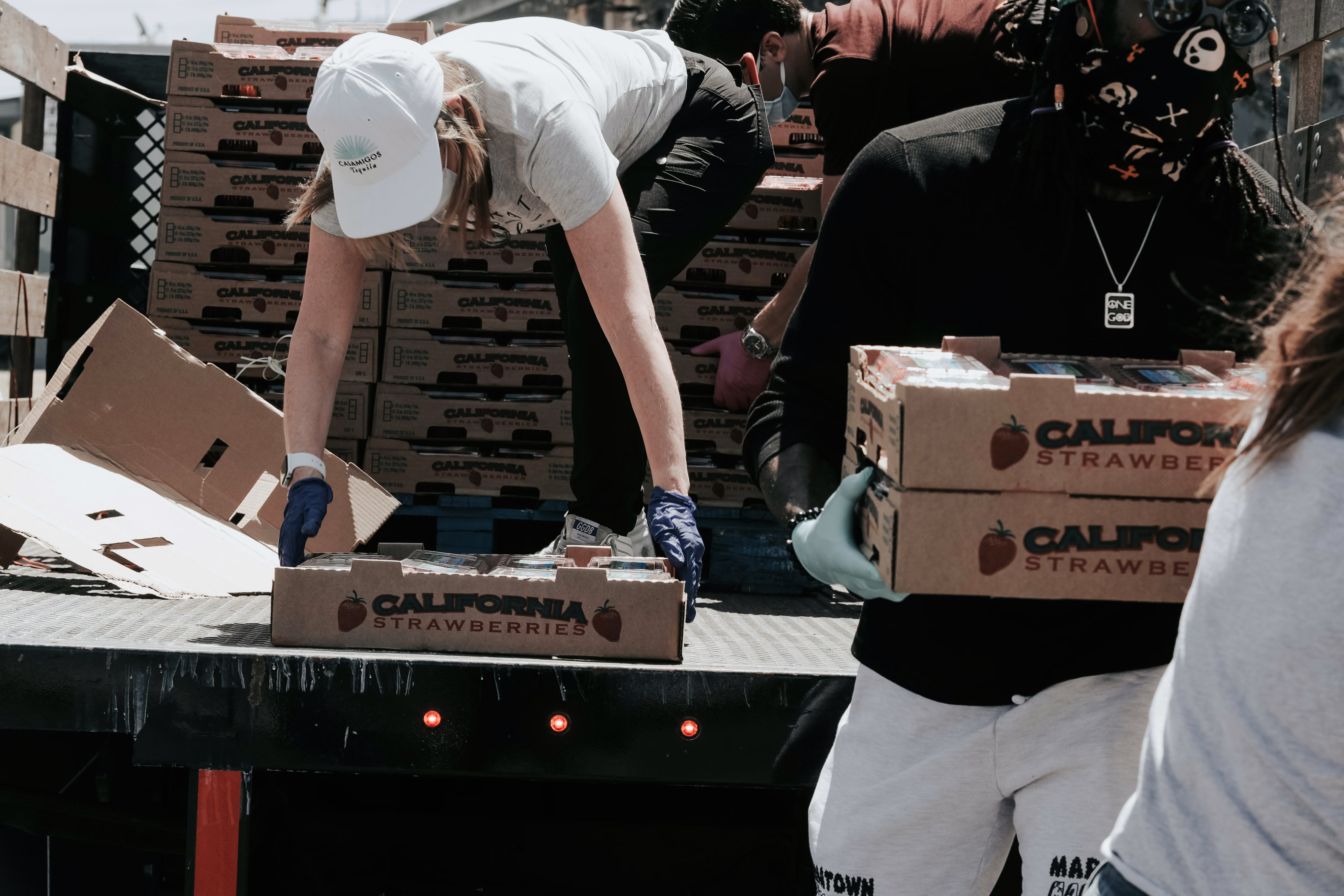 Volunteers unloading and carrying boxes labeled California strawberries from a truck wearing gloves and masks.