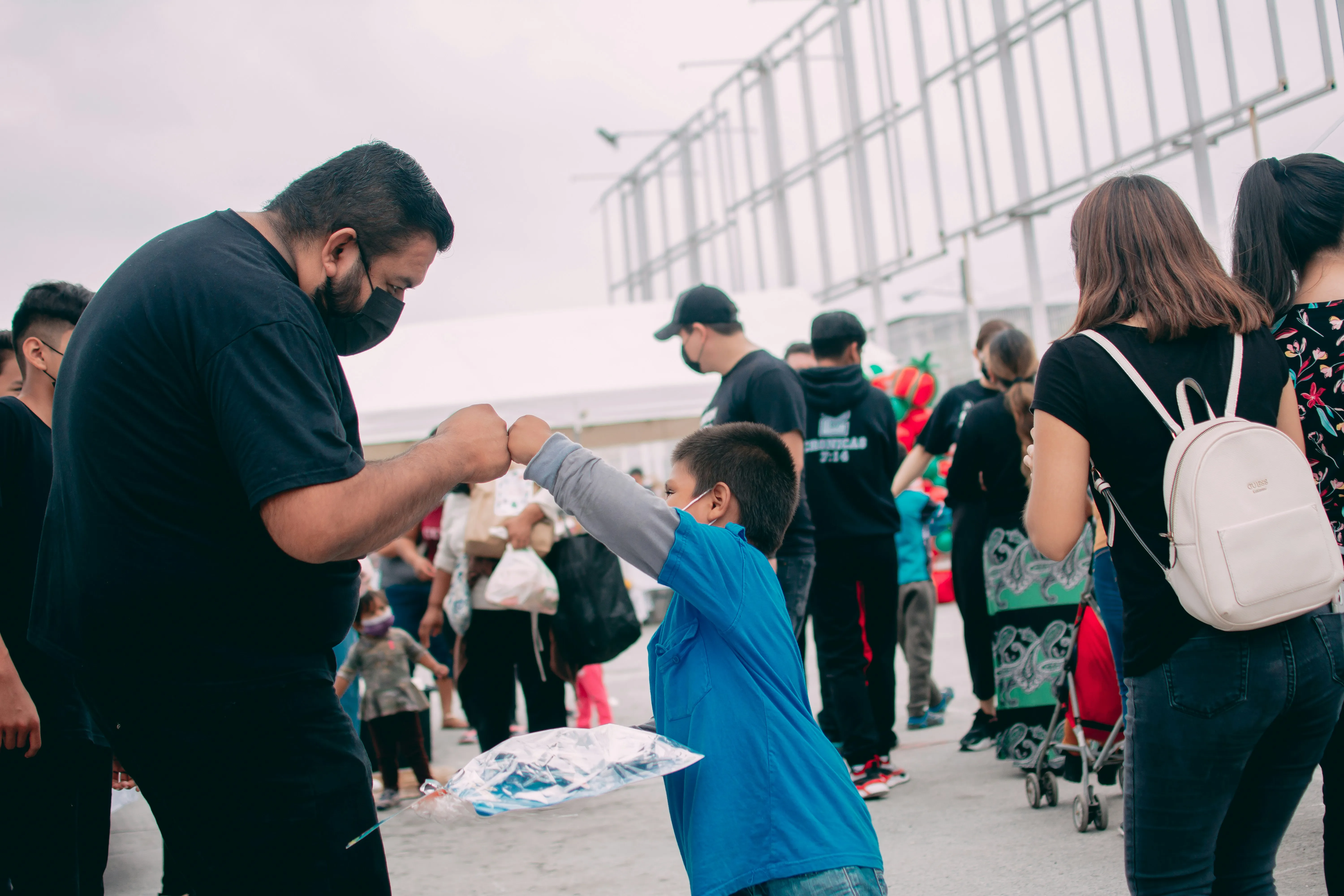 Man and boy wearing masks fist bumping outdoors in a crowd during the day.