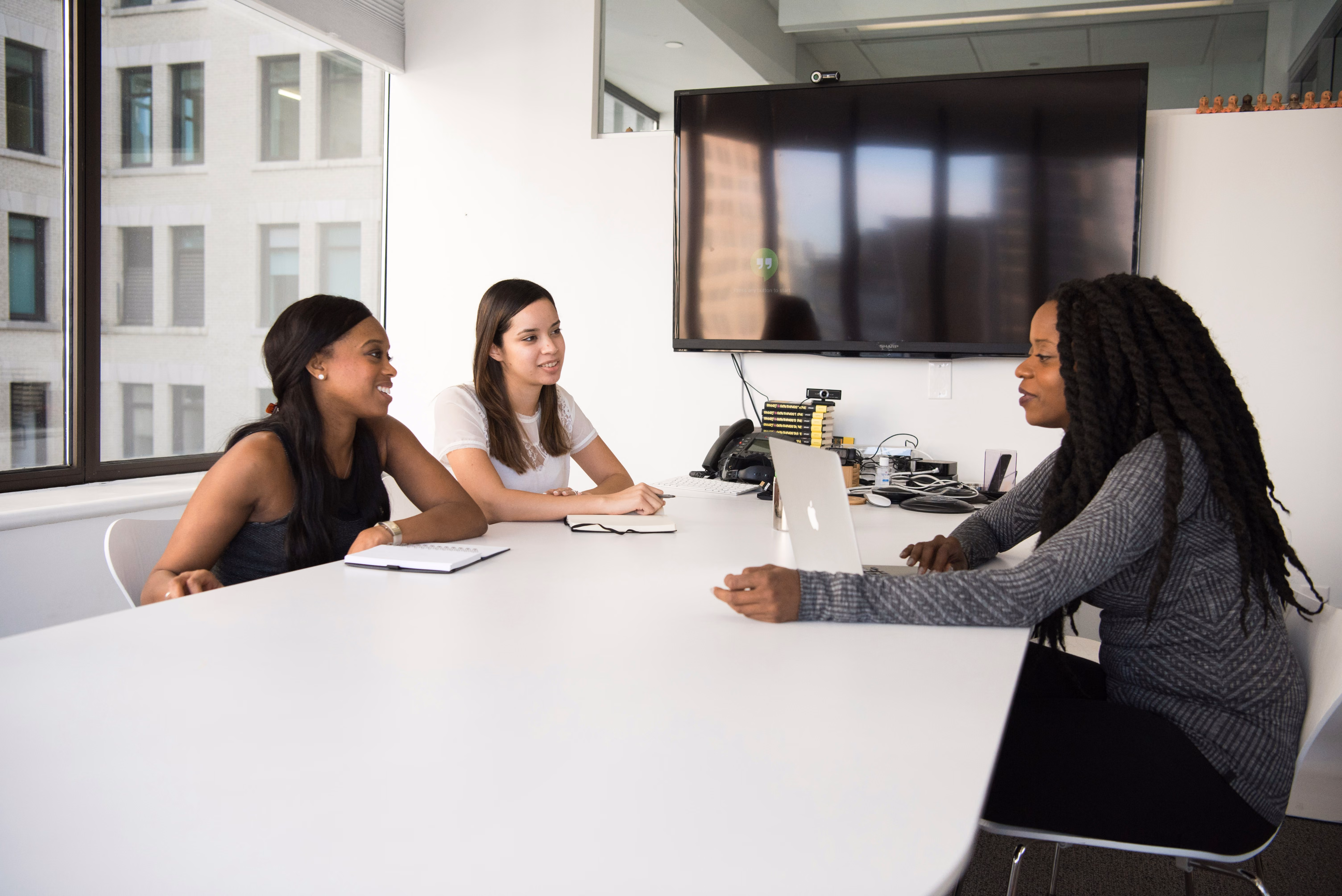 Three women having a discussion around a white conference table in an office with a laptop and notepads.