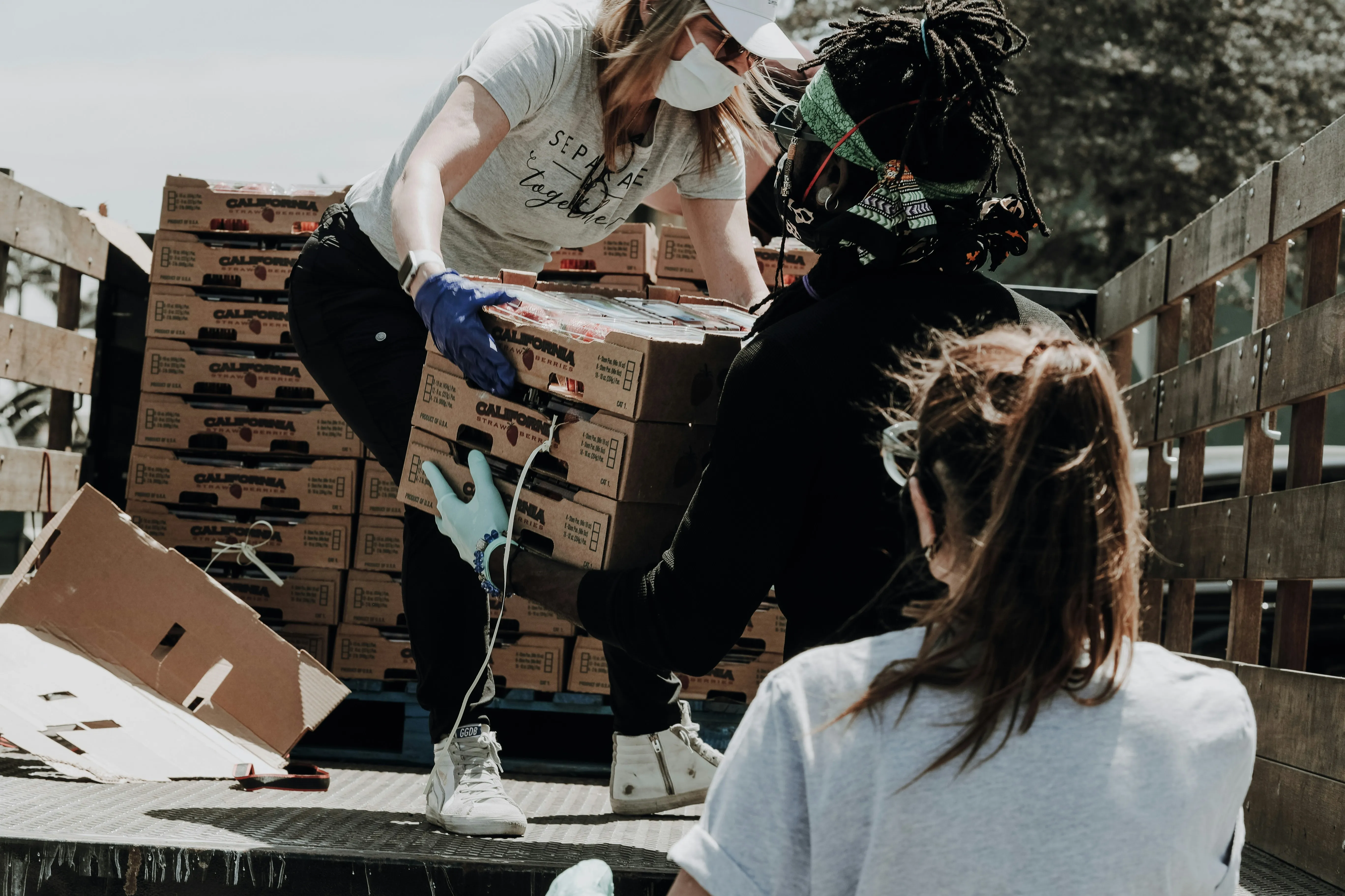 Three people wearing masks and gloves unloading boxes of California strawberries from a truck.