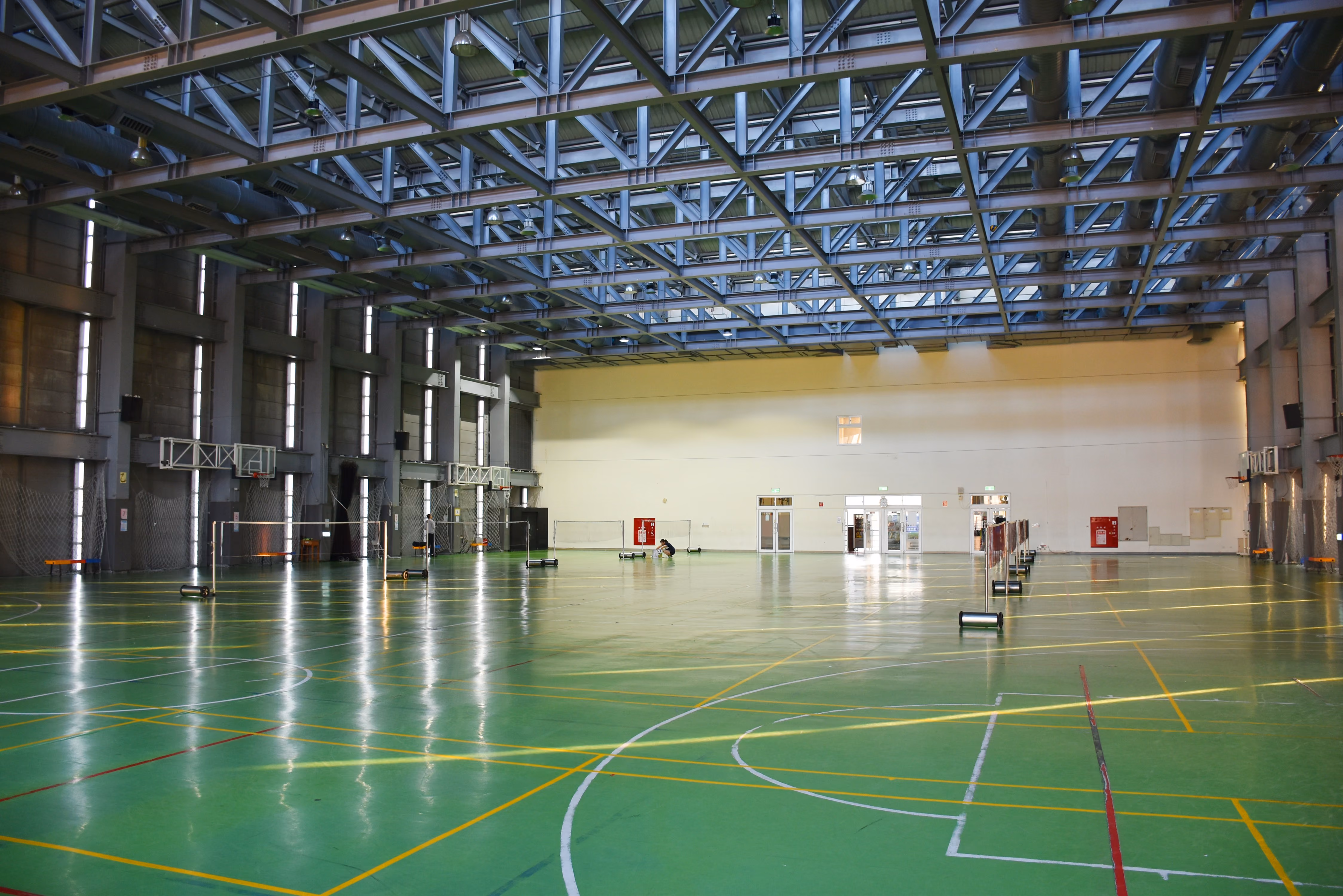 Large indoor gym with green floor, multiple badminton nets, and tall windows along the wall.