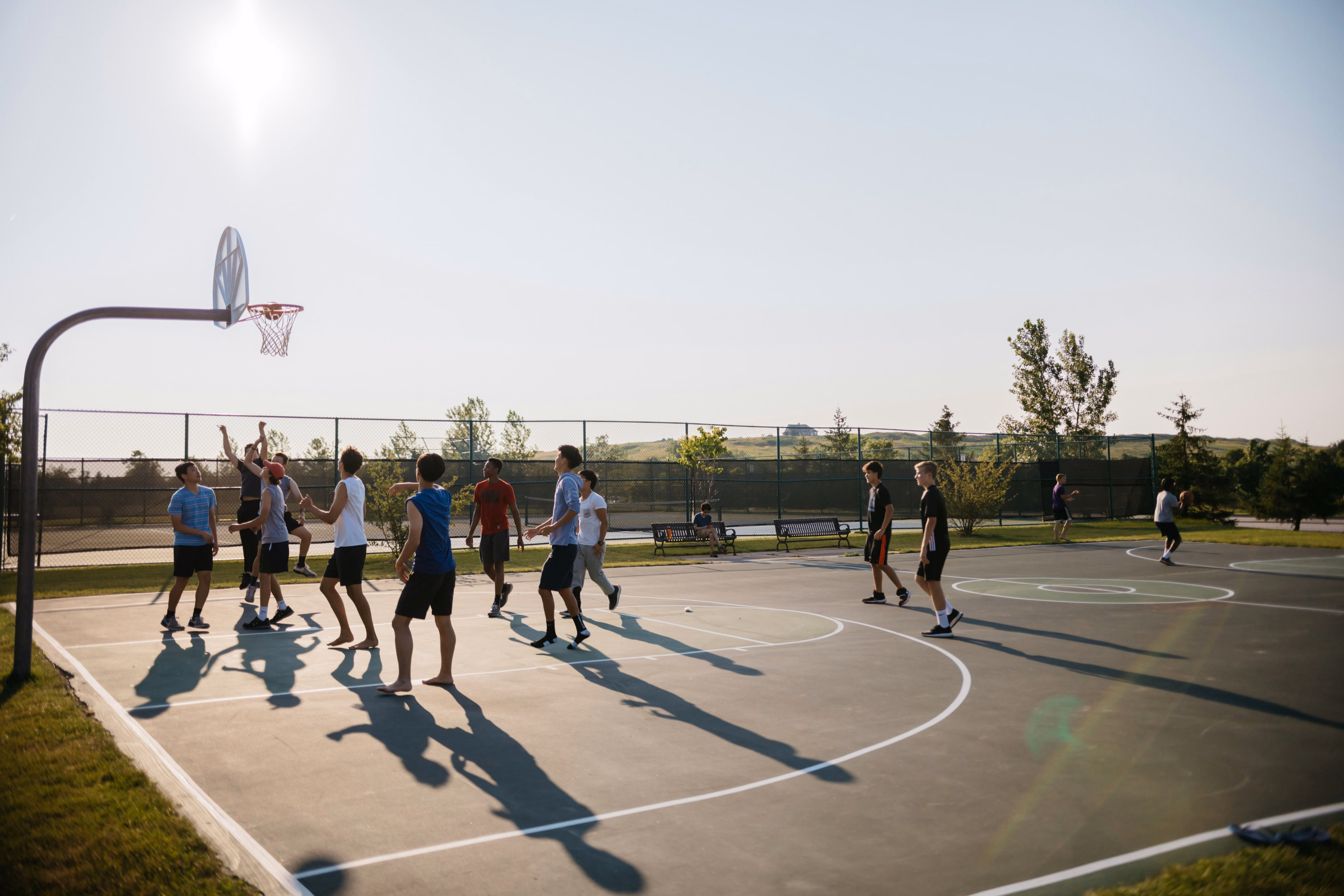 Group of young people playing basketball outdoors on a sunny court with trees and hills in the background.