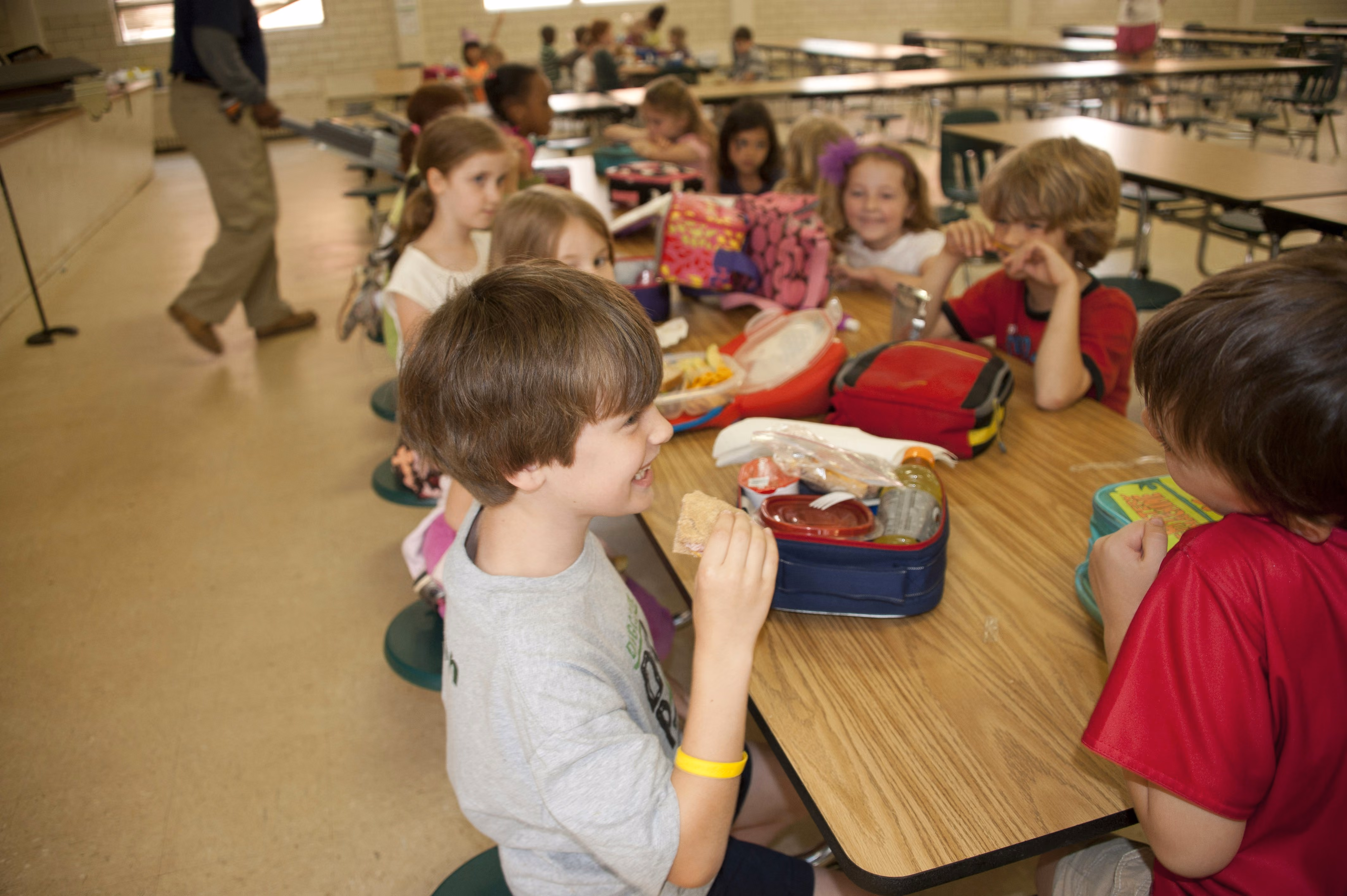 Group of children sitting at a cafeteria table eating lunch and chatting with lunchboxes in front of them.