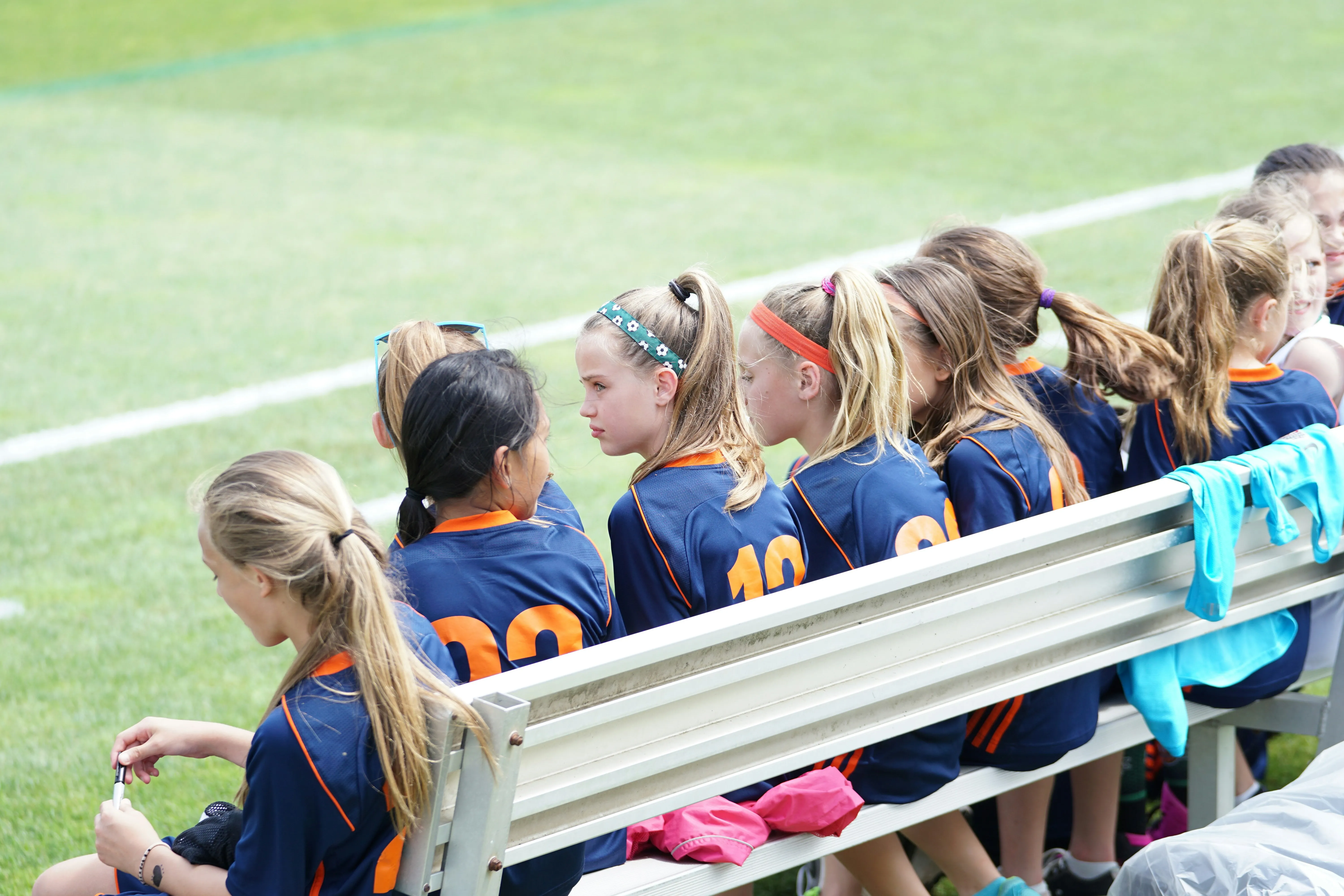 Young girls in blue and orange soccer uniforms sitting on bench by a green soccer field.