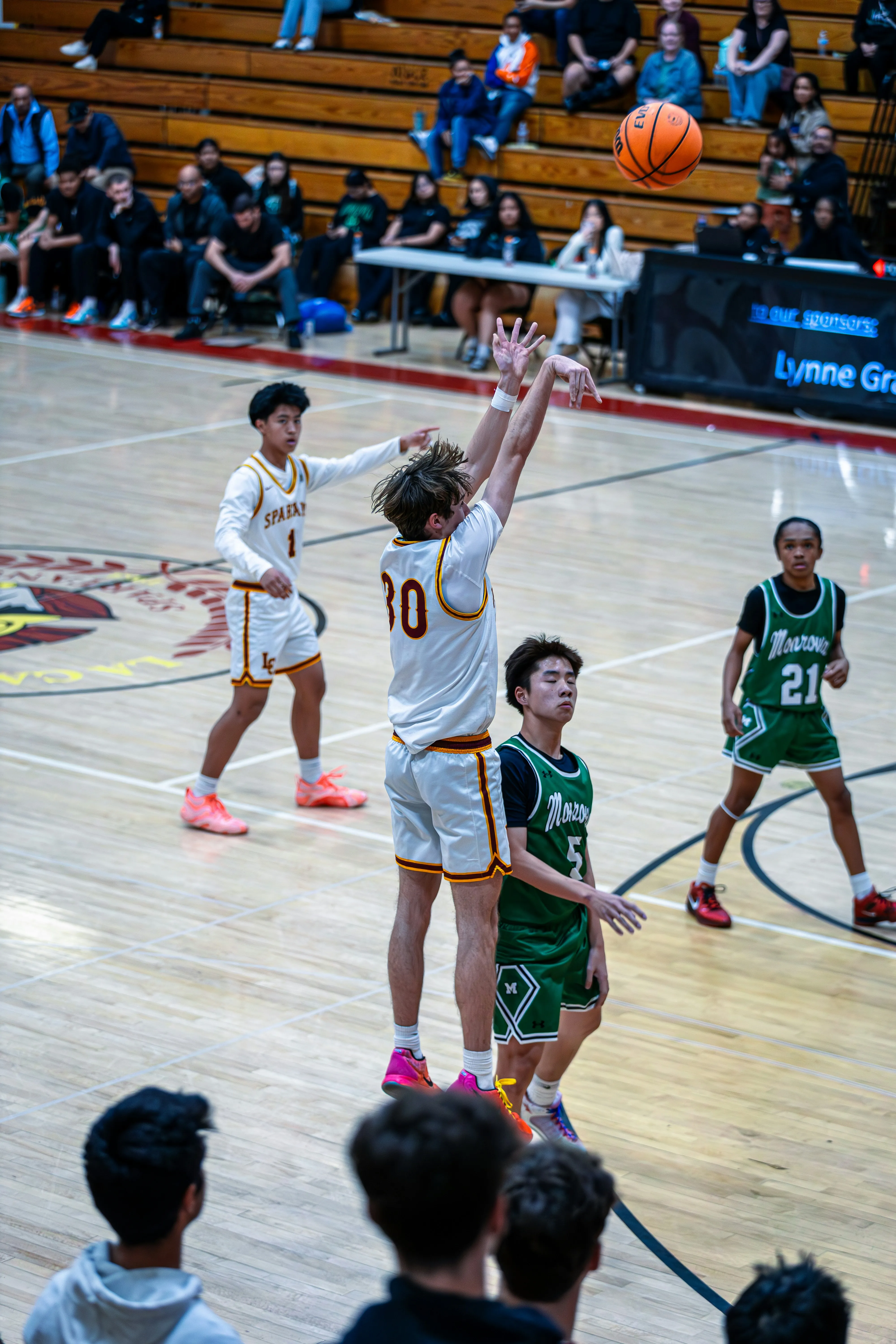 Basketball player in white uniform jumps shooting the ball while opponents in green defend on court.