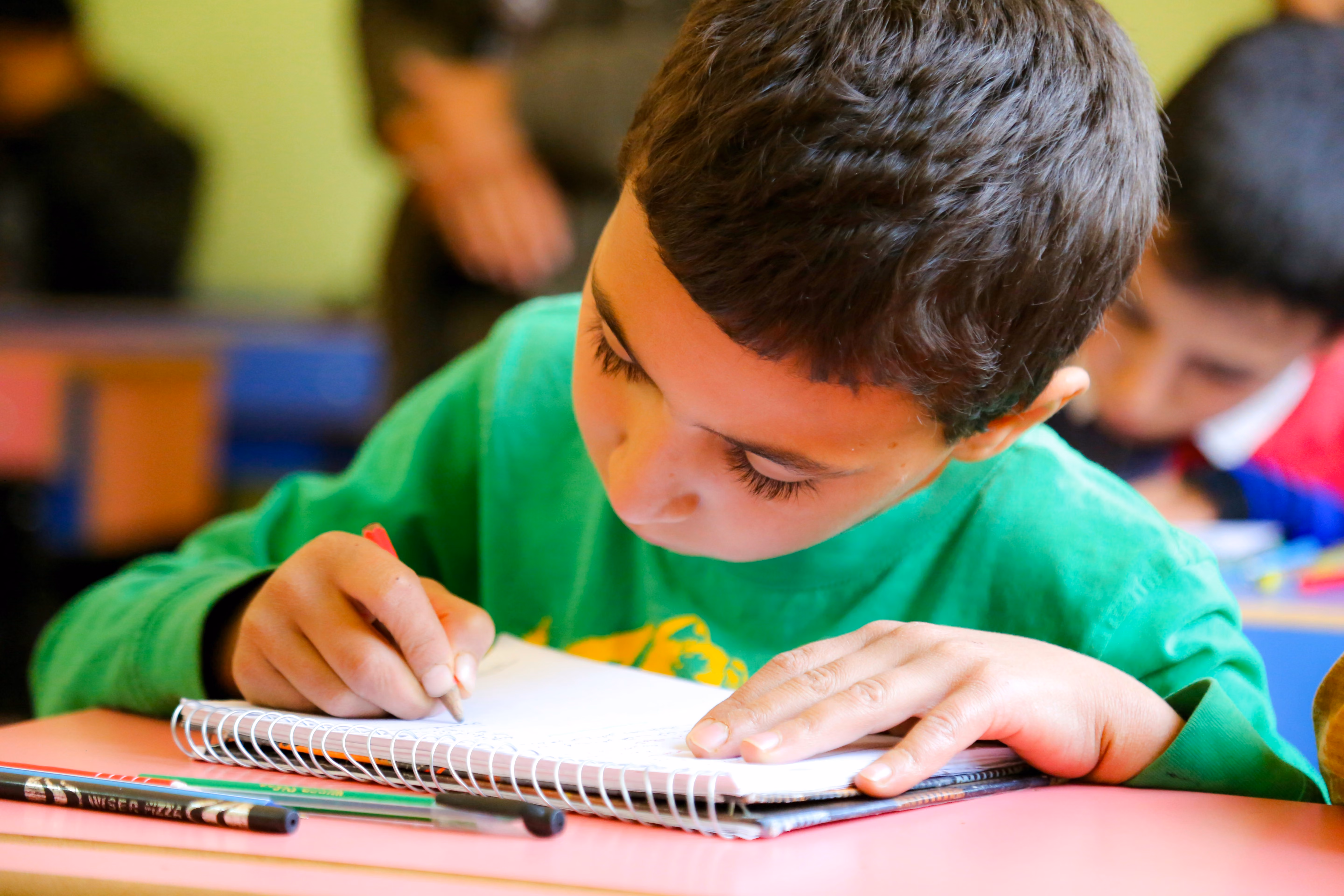 Young boy in green shirt focused on writing in a spiral notebook at a pink desk in a classroom.