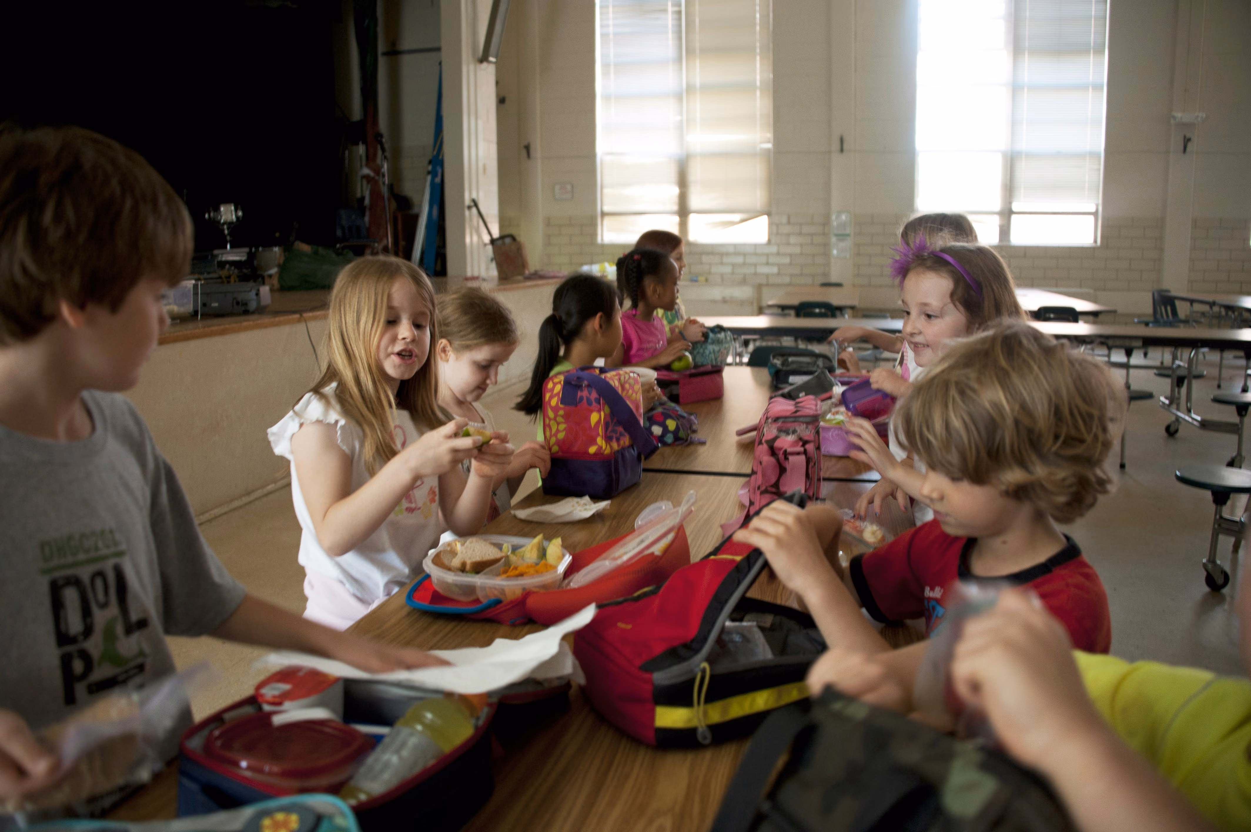 Group of young children sitting at a table in a cafeteria eating lunch and unpacking lunch boxes.