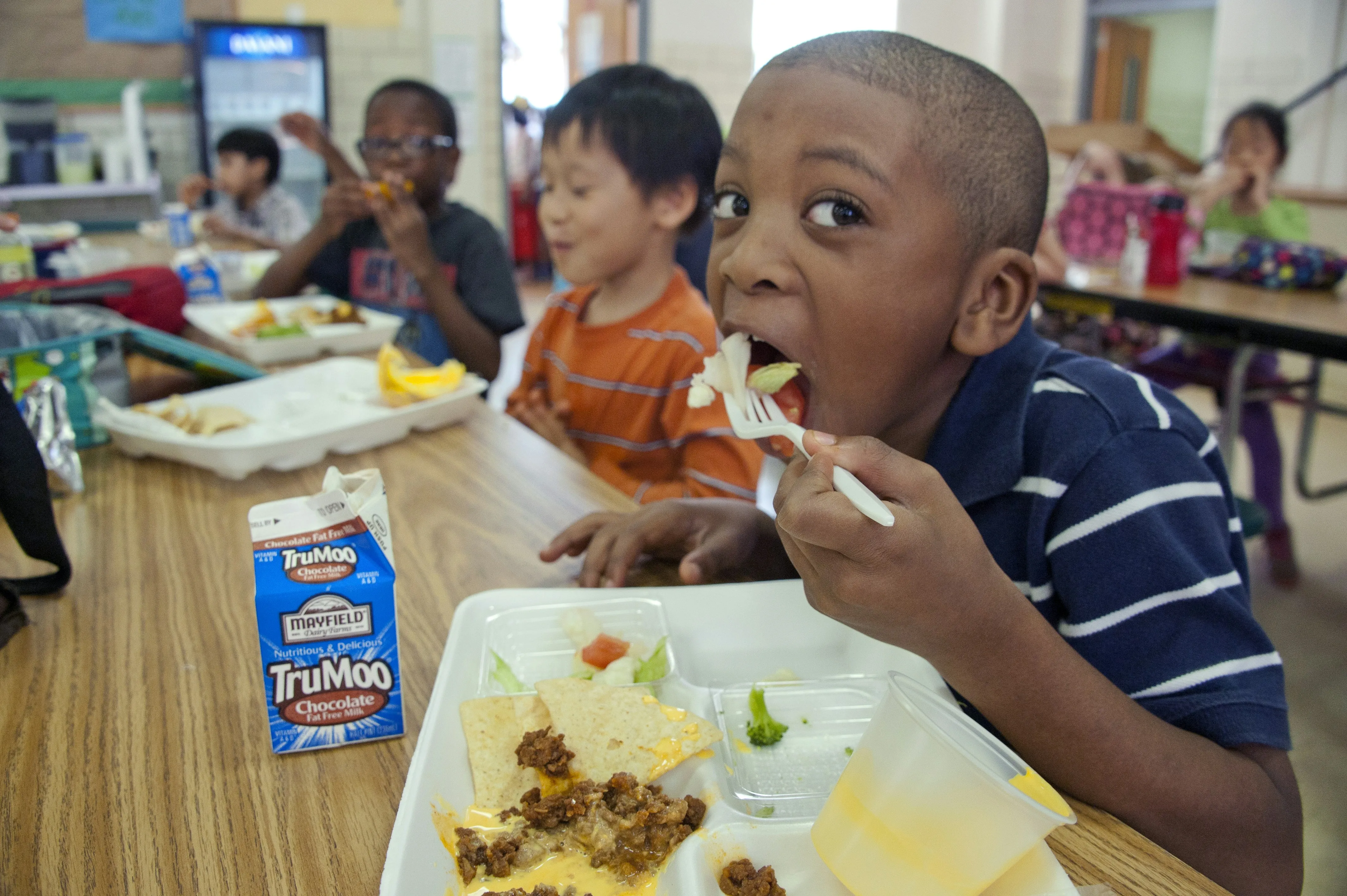 Young boy in striped shirt eating lunch with tray of food and chocolate milk carton on table.