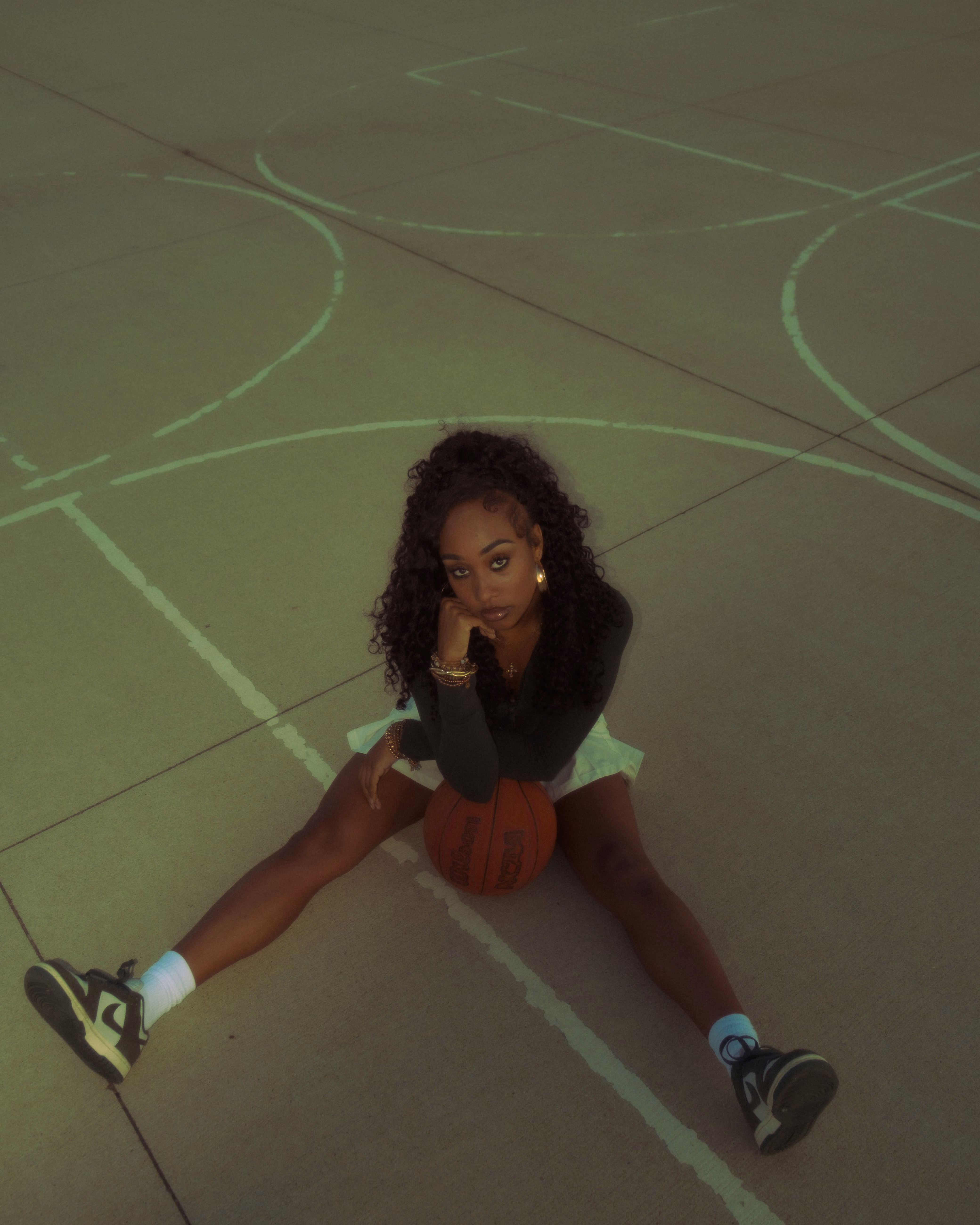 Young woman sitting on basketball court with legs apart and basketball between her legs.