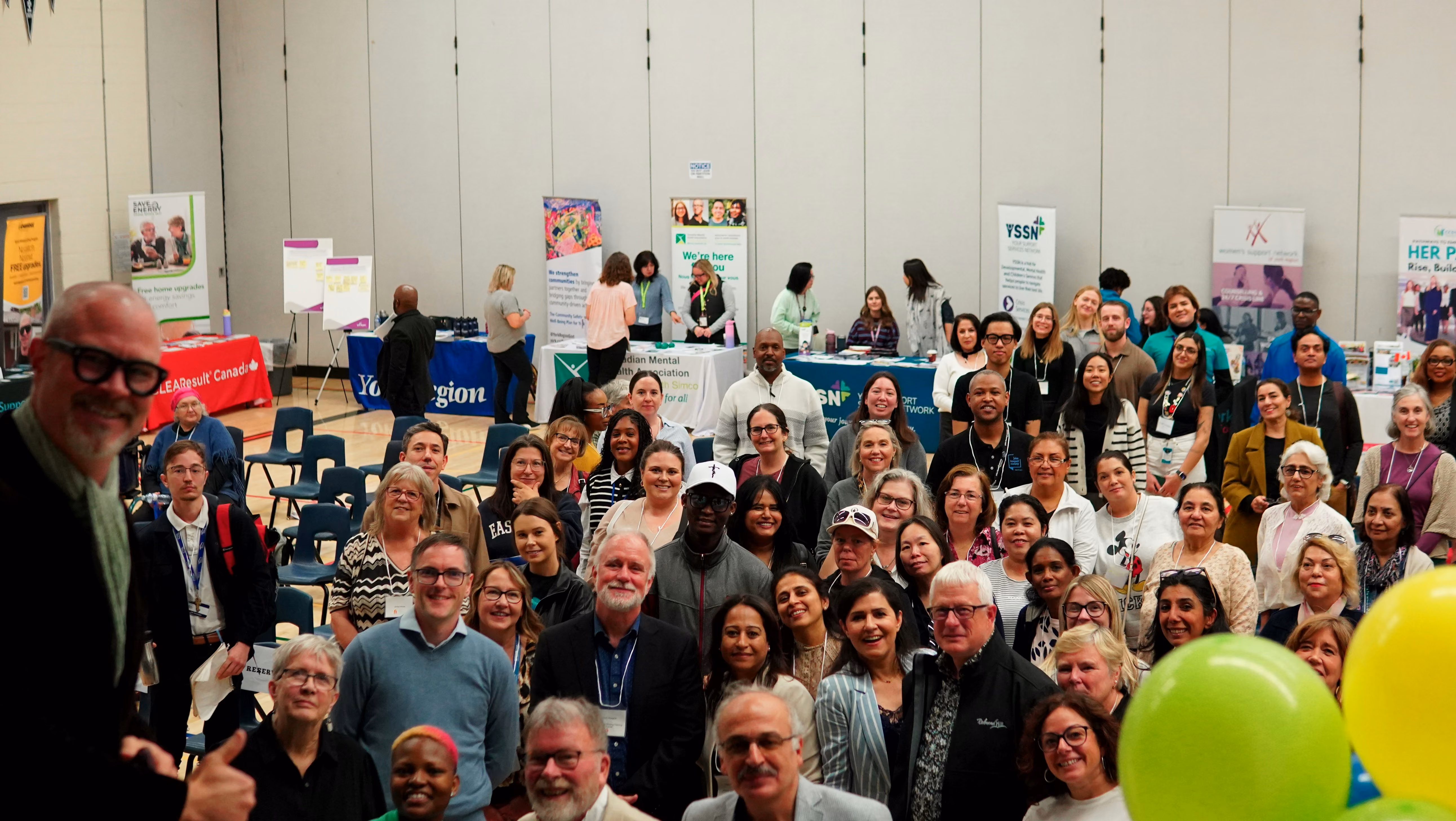 Large diverse group of people smiling at a community event in a gym with informational booths and balloons.