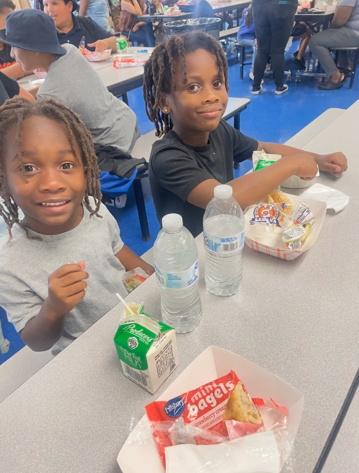 Two children with dreadlocks sitting at a cafeteria table with lunches and water bottles.
