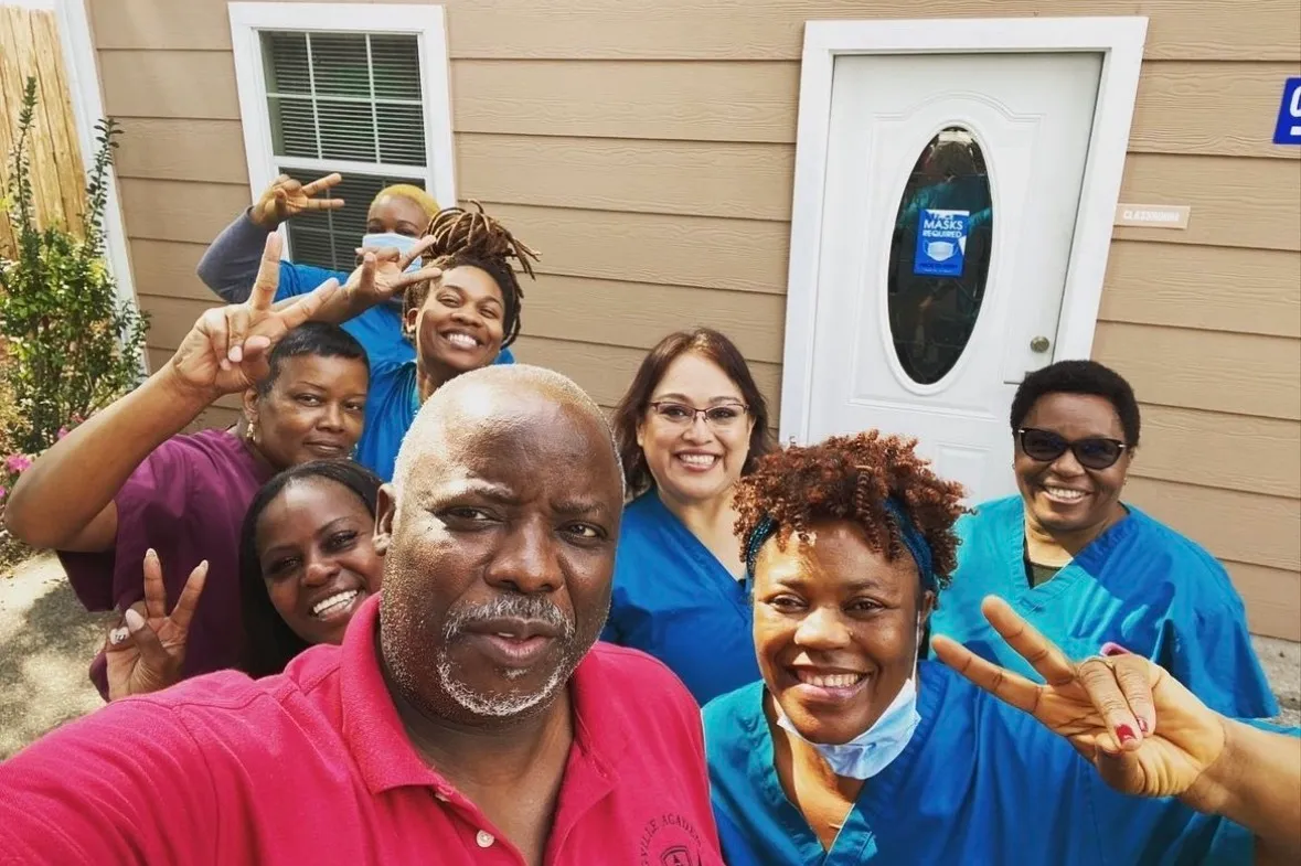 Group of smiling healthcare workers and a man taking a selfie outside a building with a mask sign on the door.