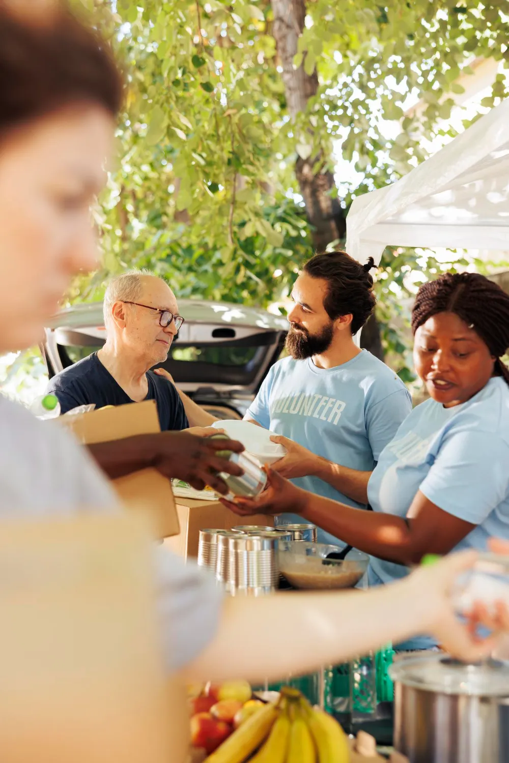 Volunteers distributing canned food and meals outdoors at a community food drive under a leafy tree.