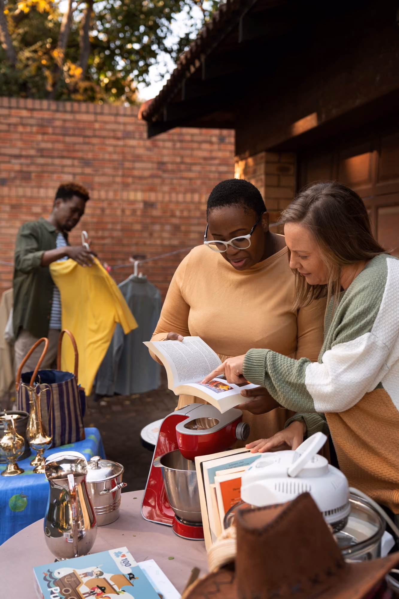 Two women looking at a book while a man browses clothes at an outdoor yard sale table with items.