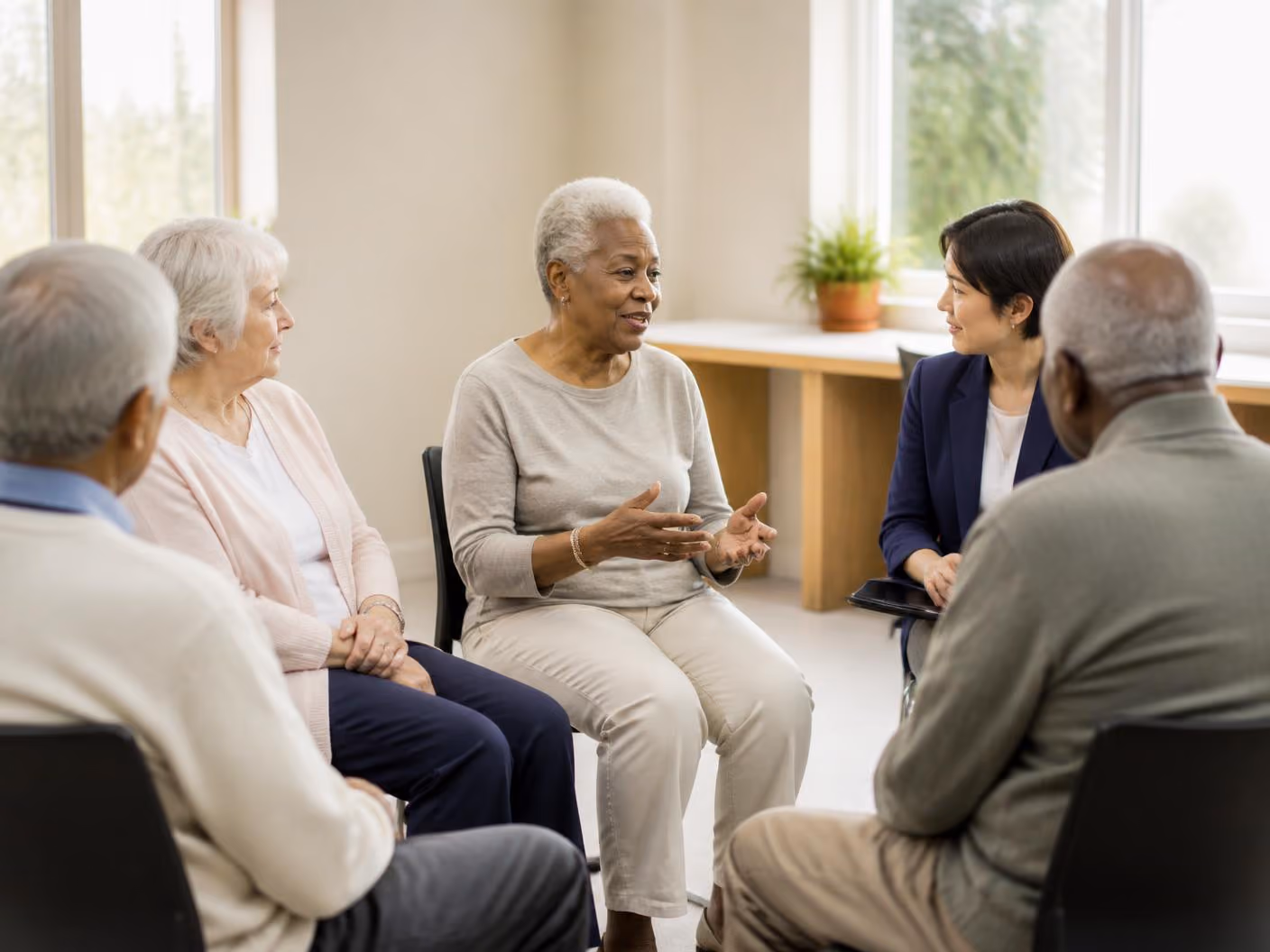 Diverse seniors sitting in a circle talking with a caregiver in a bright room with windows.