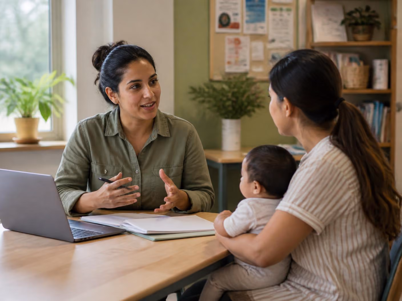 A woman advisor talks to a mother holding her baby at a desk with a laptop and documents.