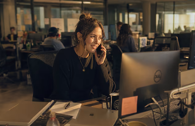 A woman in a black sweater talks on the phone at her desk with a computer in an office setting.