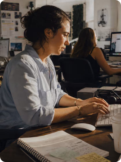 Woman with curly hair typing on keyboard at wooden desk with notebook and mouse nearby.
