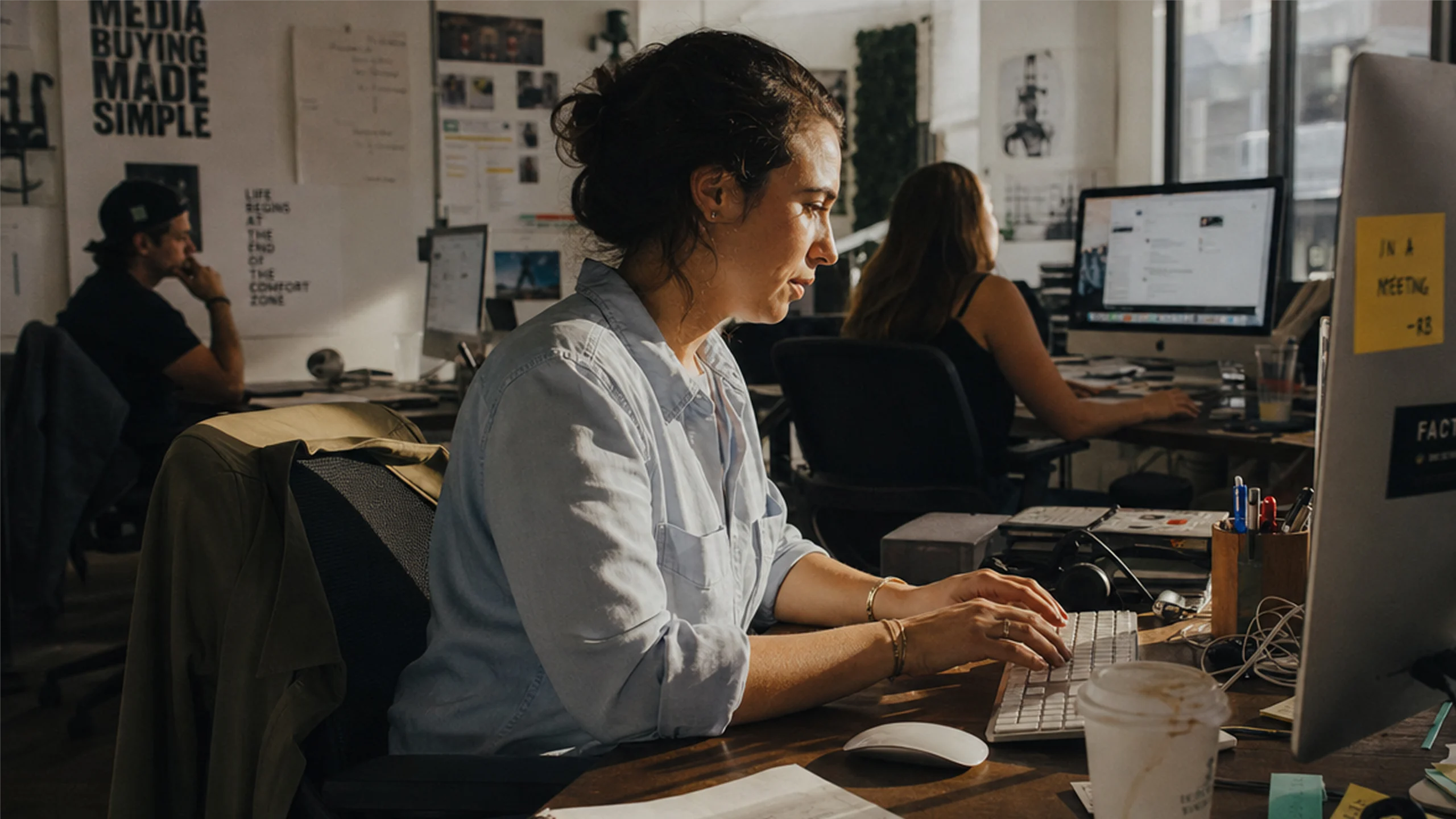 Woman in a casual shirt typing on a keyboard in a sunlit busy office with coworkers at desks.