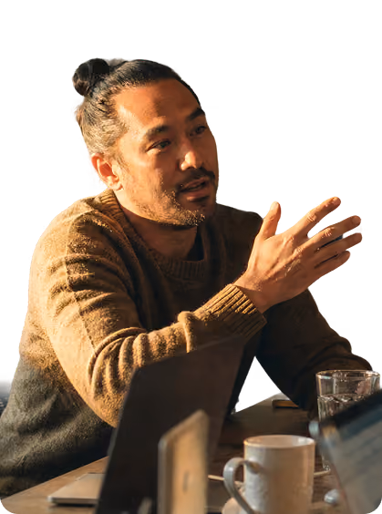 Man with man bun and brown sweater gesturing while sitting at a table with cups and laptops.