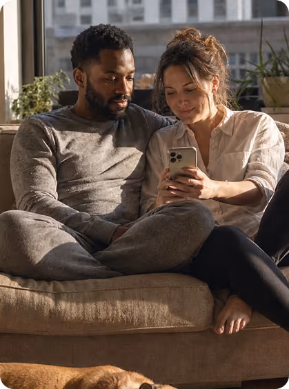 Man and woman sitting on couch together, looking at a smartphone screen and smiling.