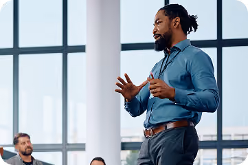 Man in blue shirt speaking and gesturing in front of seated people in a modern office with large windows.