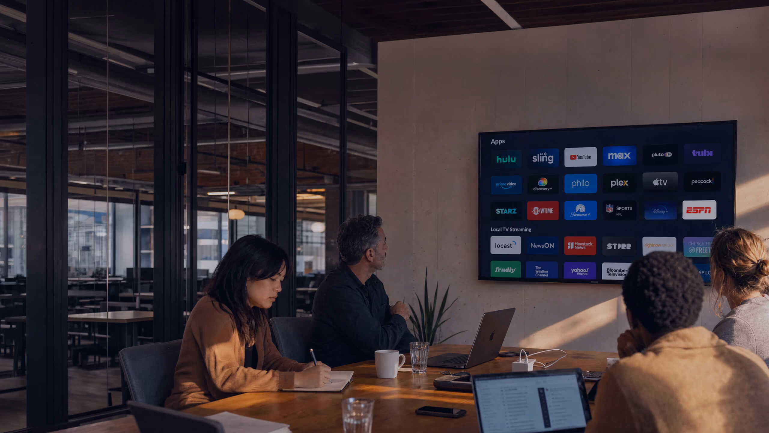 Four people are seated around a conference table in a modern office, looking at a large screen displaying various streaming app logos including Hulu, Sling, YouTube, Max, ESPN, and others.