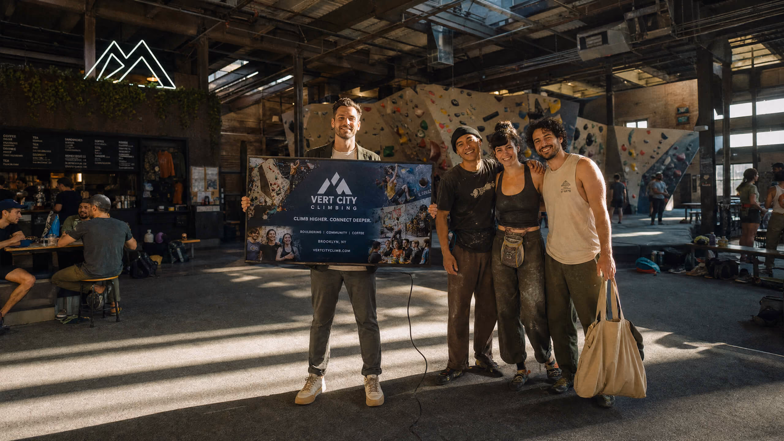 Four smiling people standing inside an indoor climbing gym with climbing walls in the background; one person is holding a sign that reads 'Vert City Climbing.'