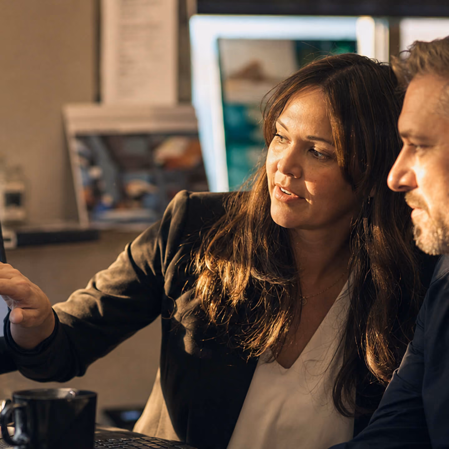A woman with long brown hair and a black blazer is explaining something to a man with light hair and facial hair as they look at a screen together in an indoor setting.