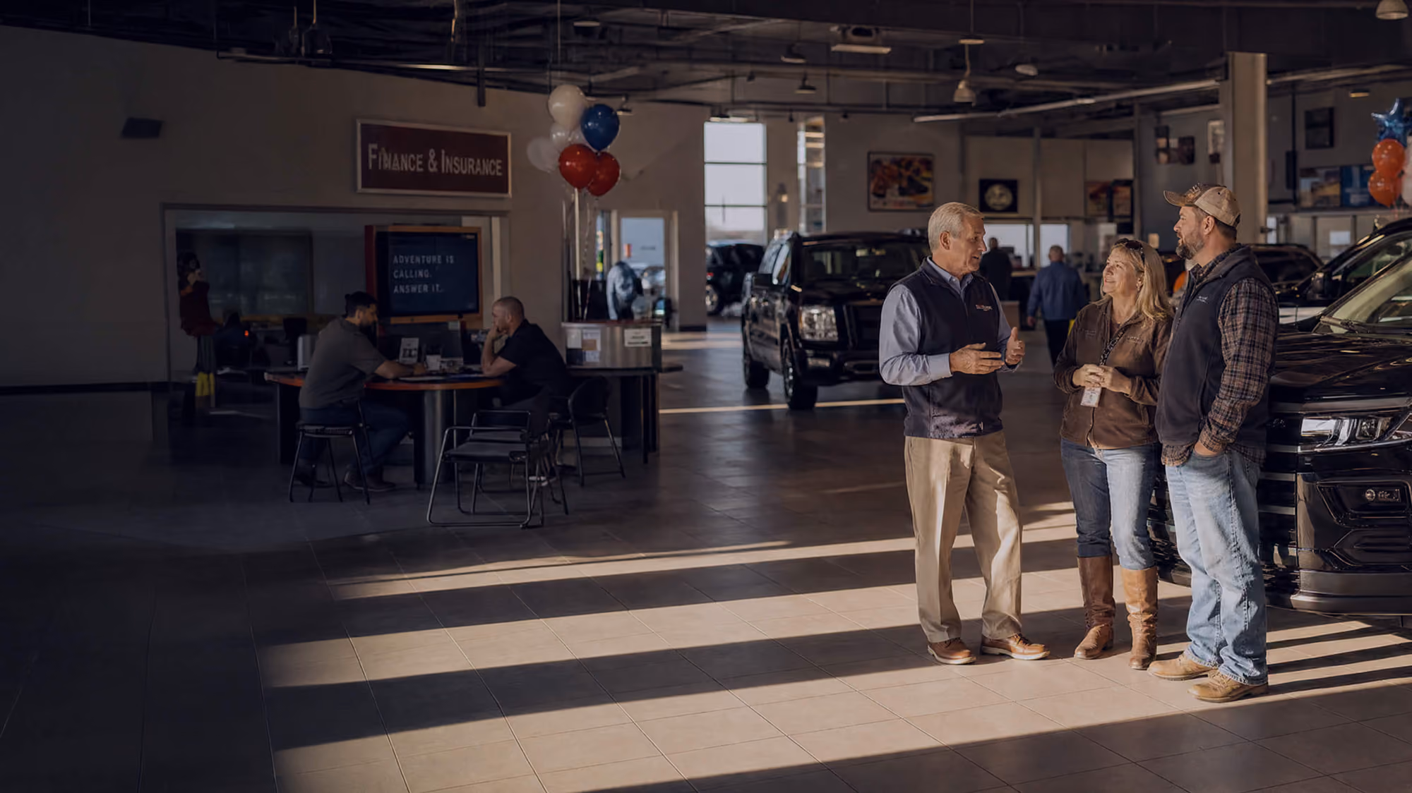 Inside a car dealership, a salesperson talks with a couple standing beside a black vehicle, while two other people sit at a table in the background near a sign that reads 'Finance & Insurance.'