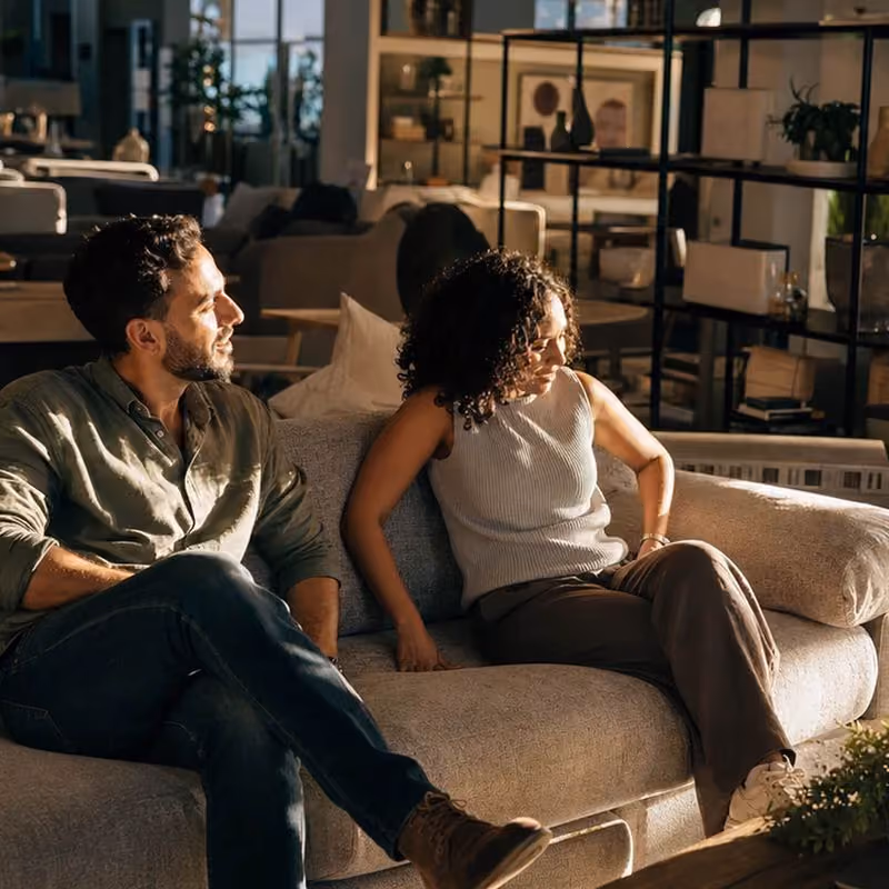 A man and a woman sitting on a beige couch in a warmly lit living room. The man wears a green shirt and jeans, looking to his left, while the woman in a sleeveless white top and brown pants turns slightly away.