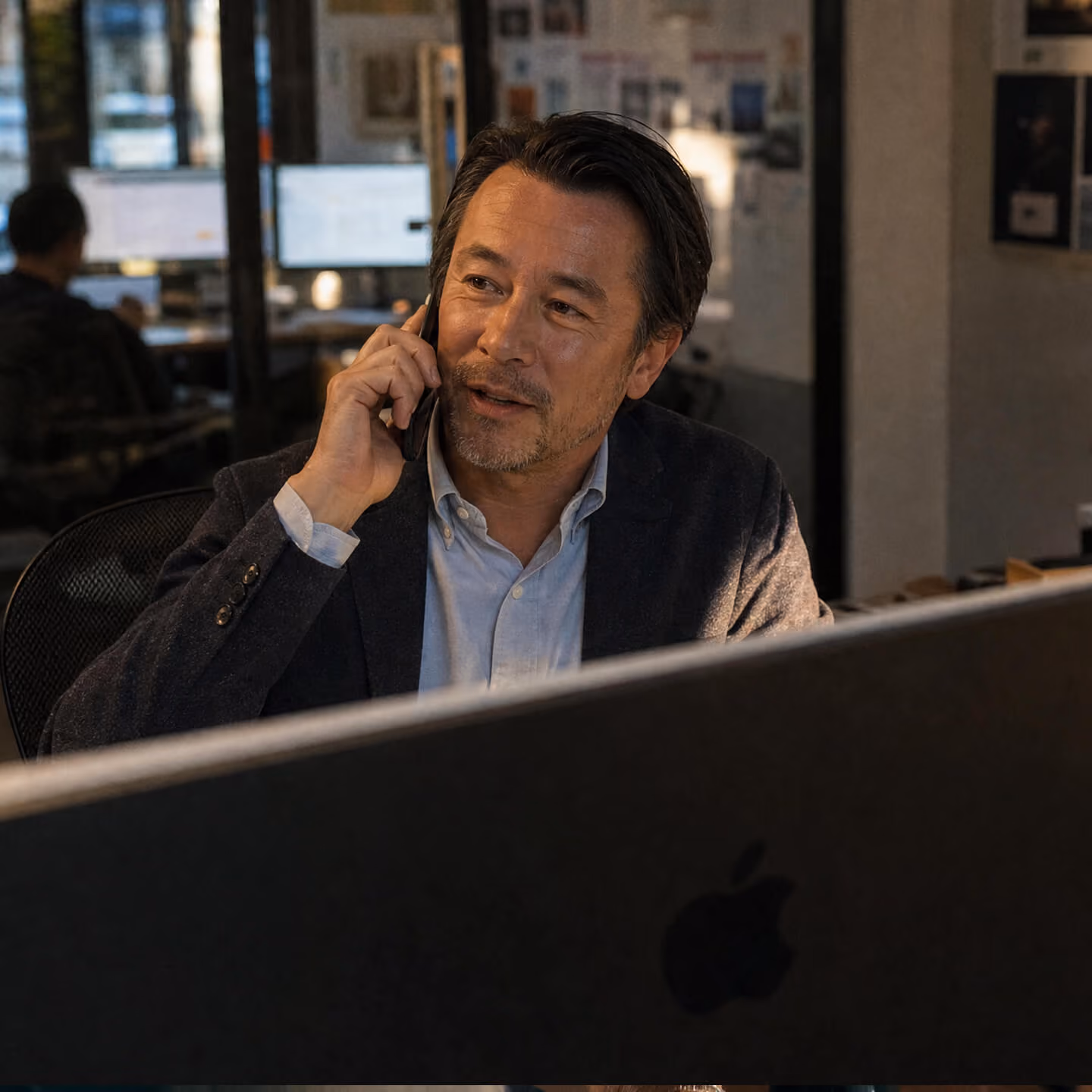 Middle-aged man with short dark hair and light beard talking on a smartphone while sitting at a desk with a large Apple computer monitor in an office.