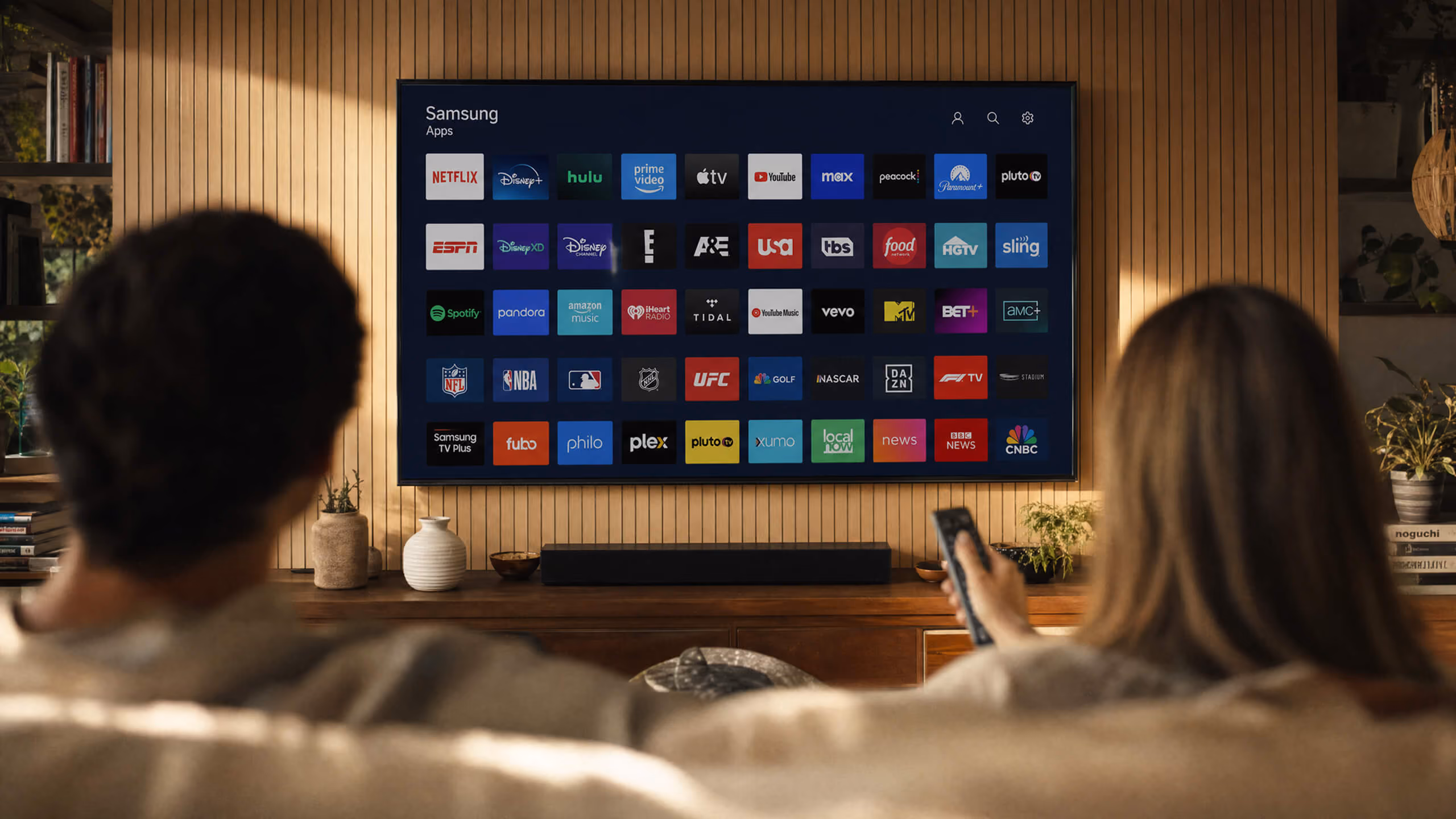 A couple sitting on a couch watching a smart TV displaying a Samsung apps menu with streaming services like Netflix, Disney+, Hulu, Prime Video, Apple TV, YouTube, and others on a wooden-paneled wall.