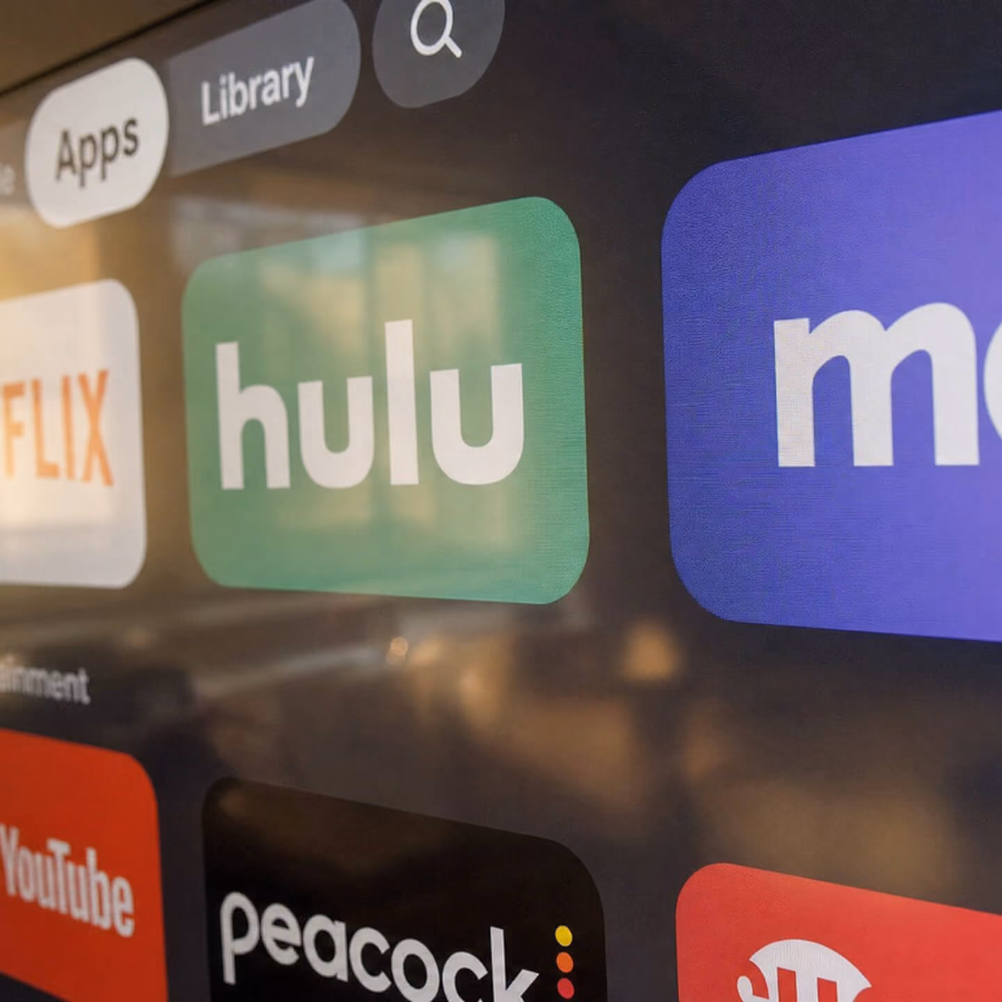 Close-up of a smart TV screen showing app icons including Hulu, Netflix, YouTube, and Peacock, with a search icon and tabs labeled Apps and Library.
