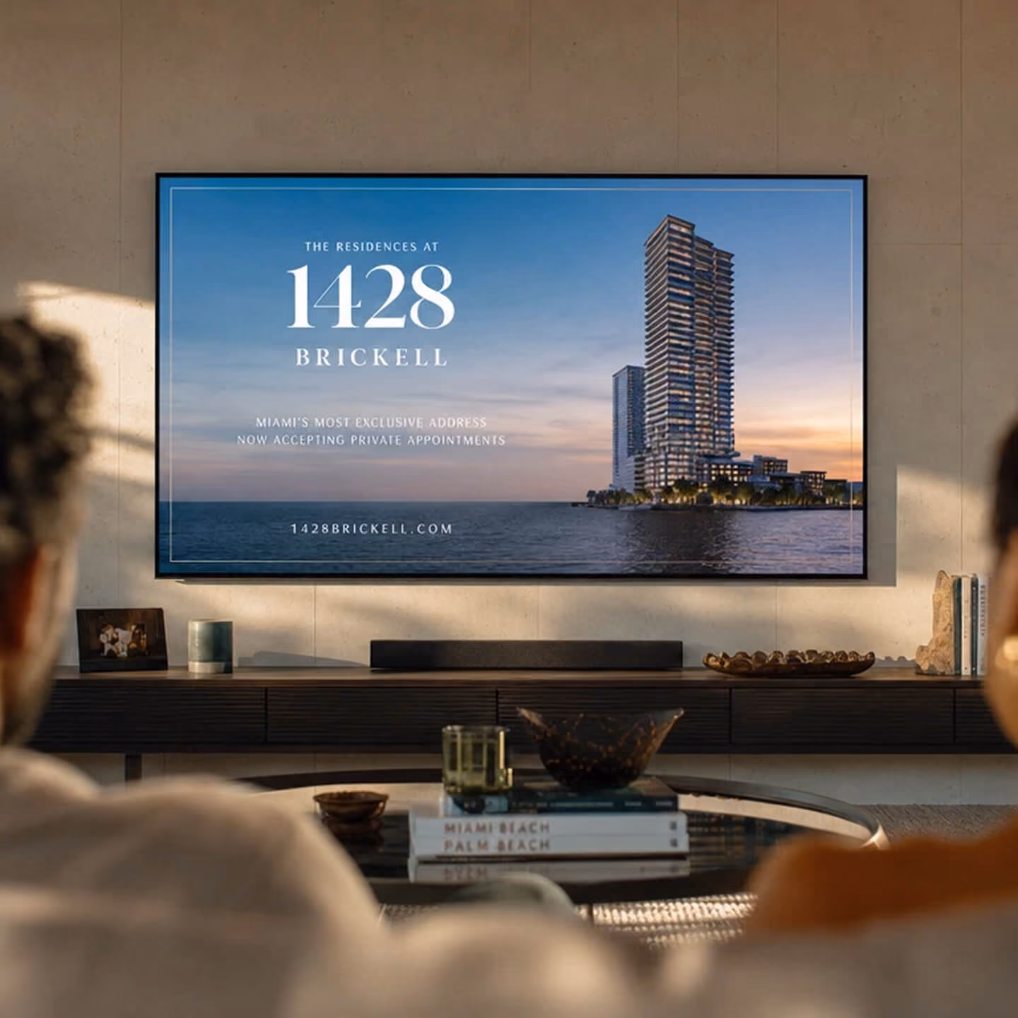 A couple sitting indoors watching a large wall-mounted screen showing an advertisement for 1428 Brickell, a residential building complex by the water at sunset in Miami, with the text 'The Residences at 1428 Brickell, Miami's Most Exclusive Address, Now Accepting Private Appointments, 1428brickell.com'.