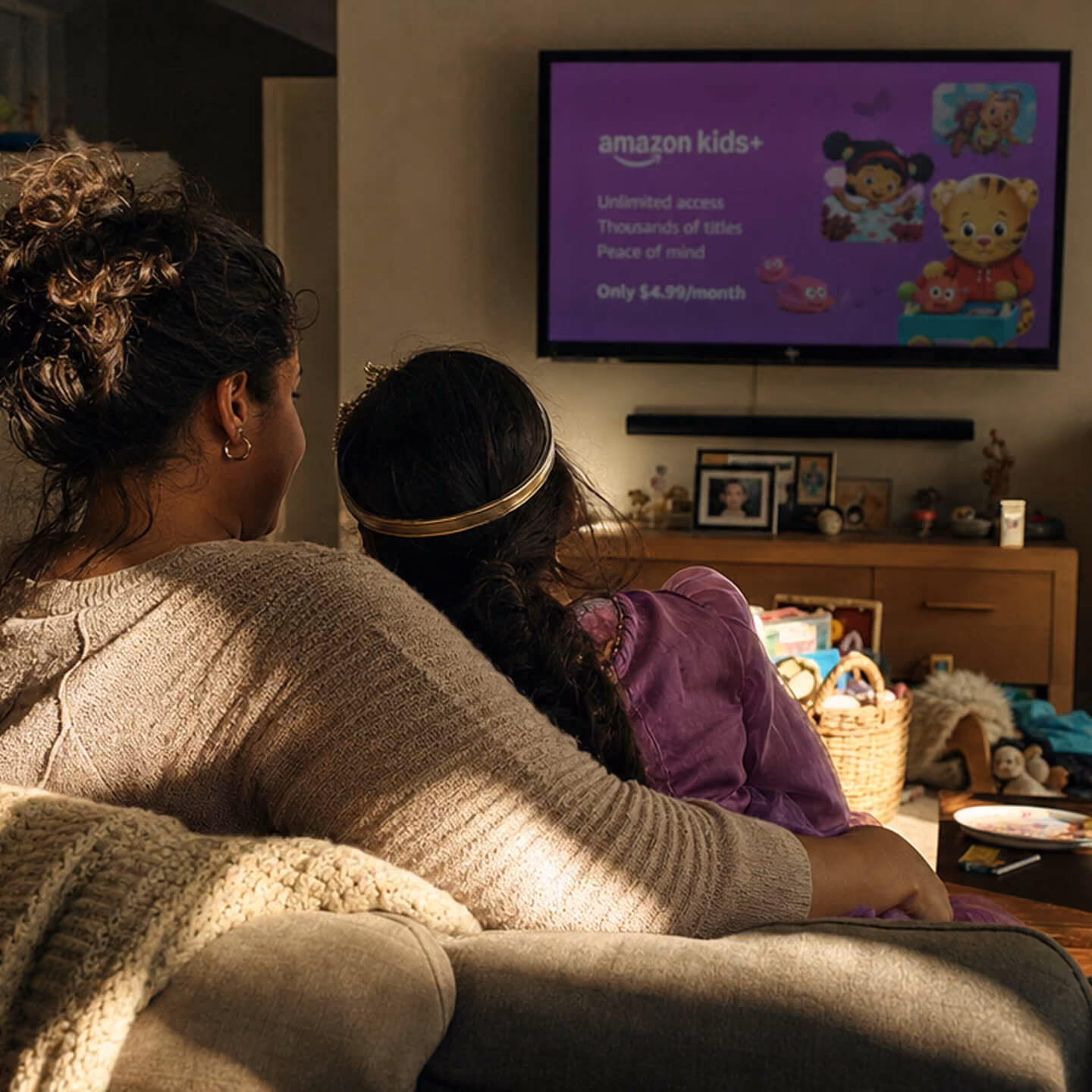 A woman and a young girl sitting closely on a couch, watching a wall-mounted TV screen displaying an Amazon Kids+ advertisement with characters on a purple background.