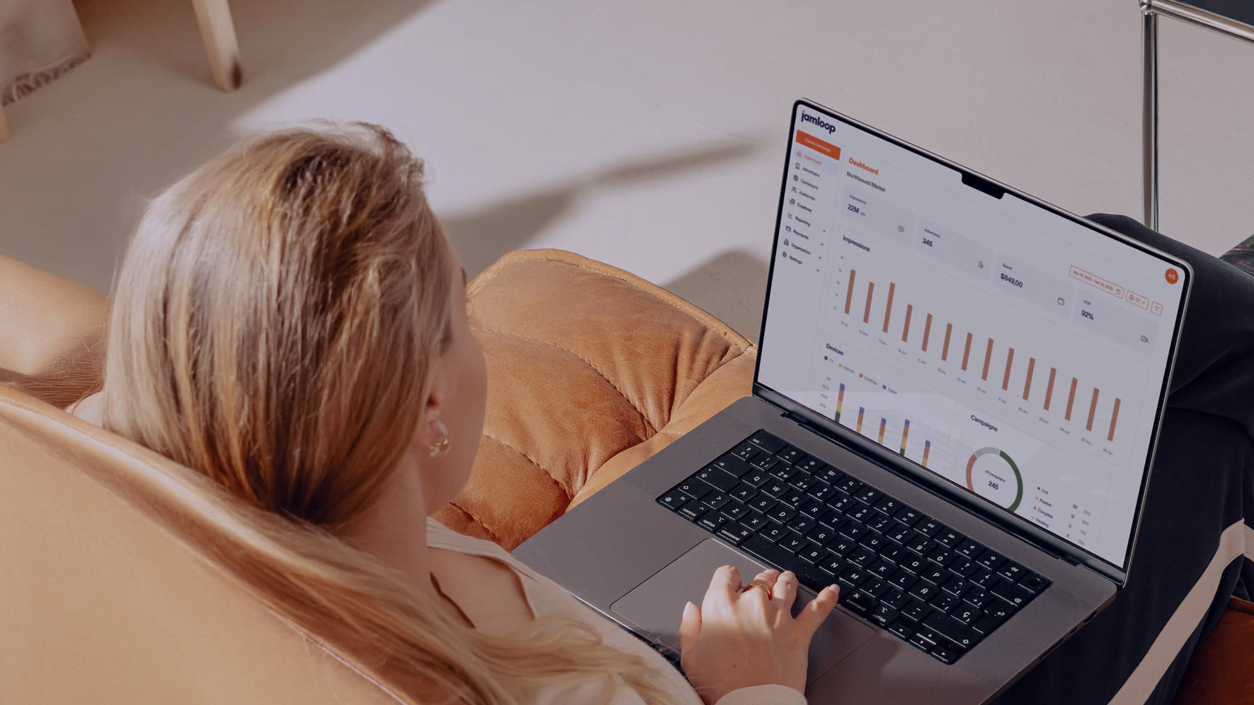 Person with long blonde hair sitting on a brown leather chair using a laptop that displays a performance dashboard with bar charts and graphs.
