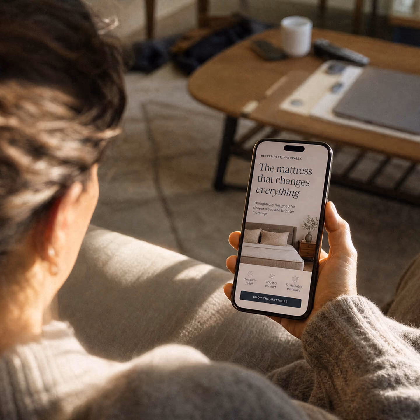 Person holding a smartphone showing a mattress advertisement with the text 'The mattress that changes everything' and icons for pressure relief, cooling comfort, and sustainable materials, with a button to shop the mattress.