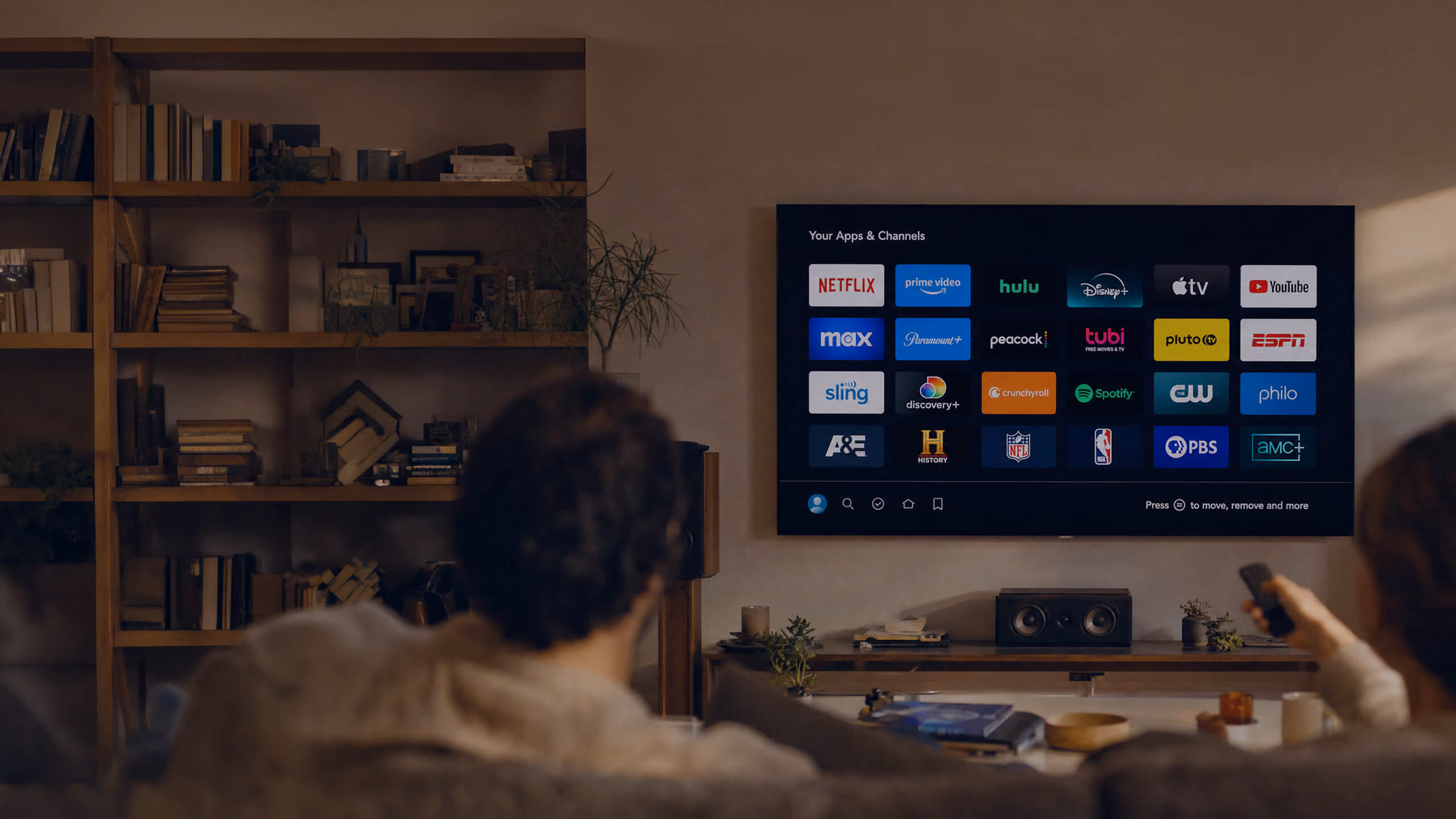 A couple sitting on a couch watching a smart TV displaying a streaming app menu with services like Netflix, Disney+, Hulu, and YouTube in a cozy living room with a bookshelf in the background.