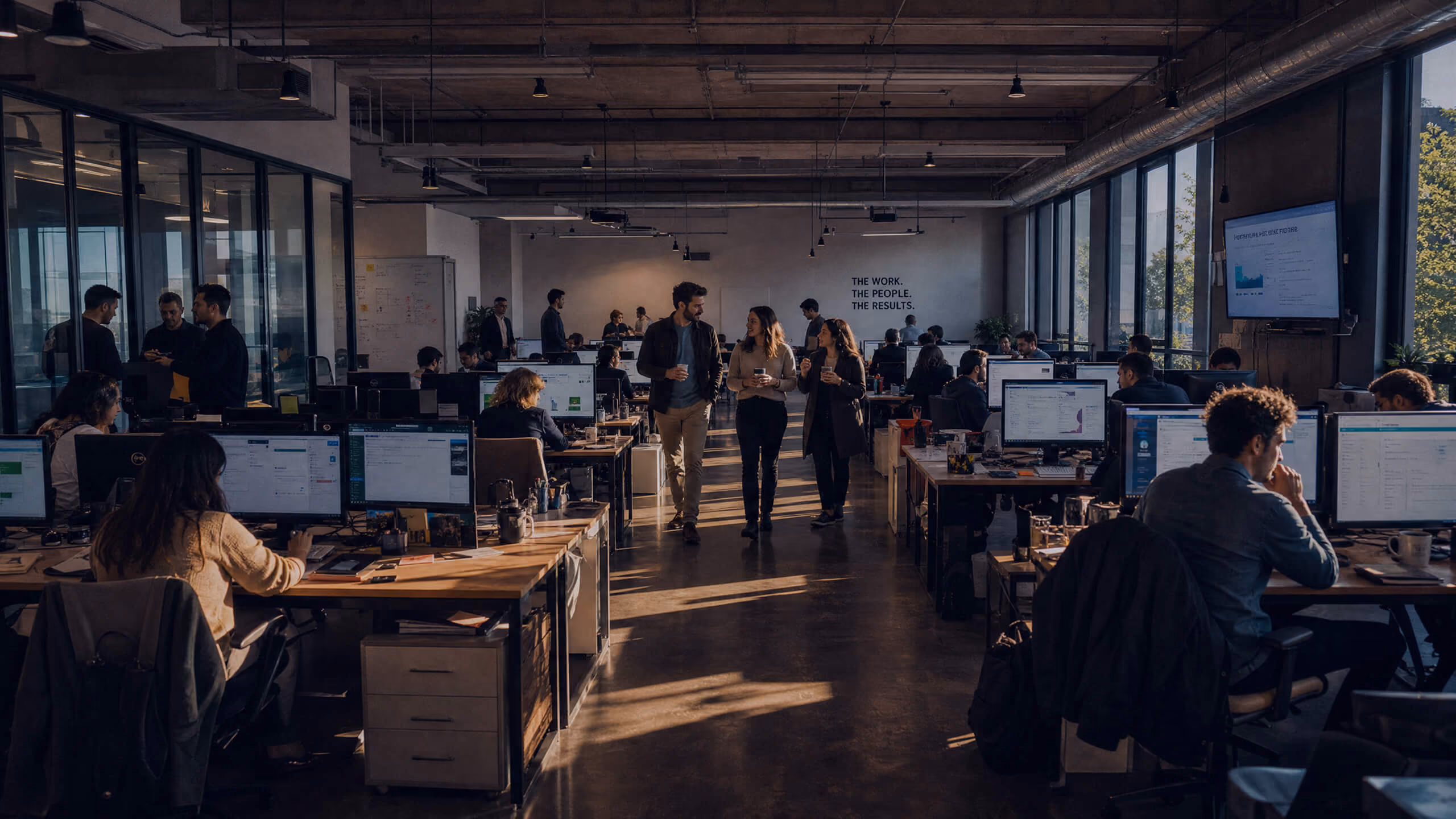 Open office space with rows of desks and computer monitors where people are working and three colleagues walking and talking in the center aisle under overhead lighting and large windows on one side.